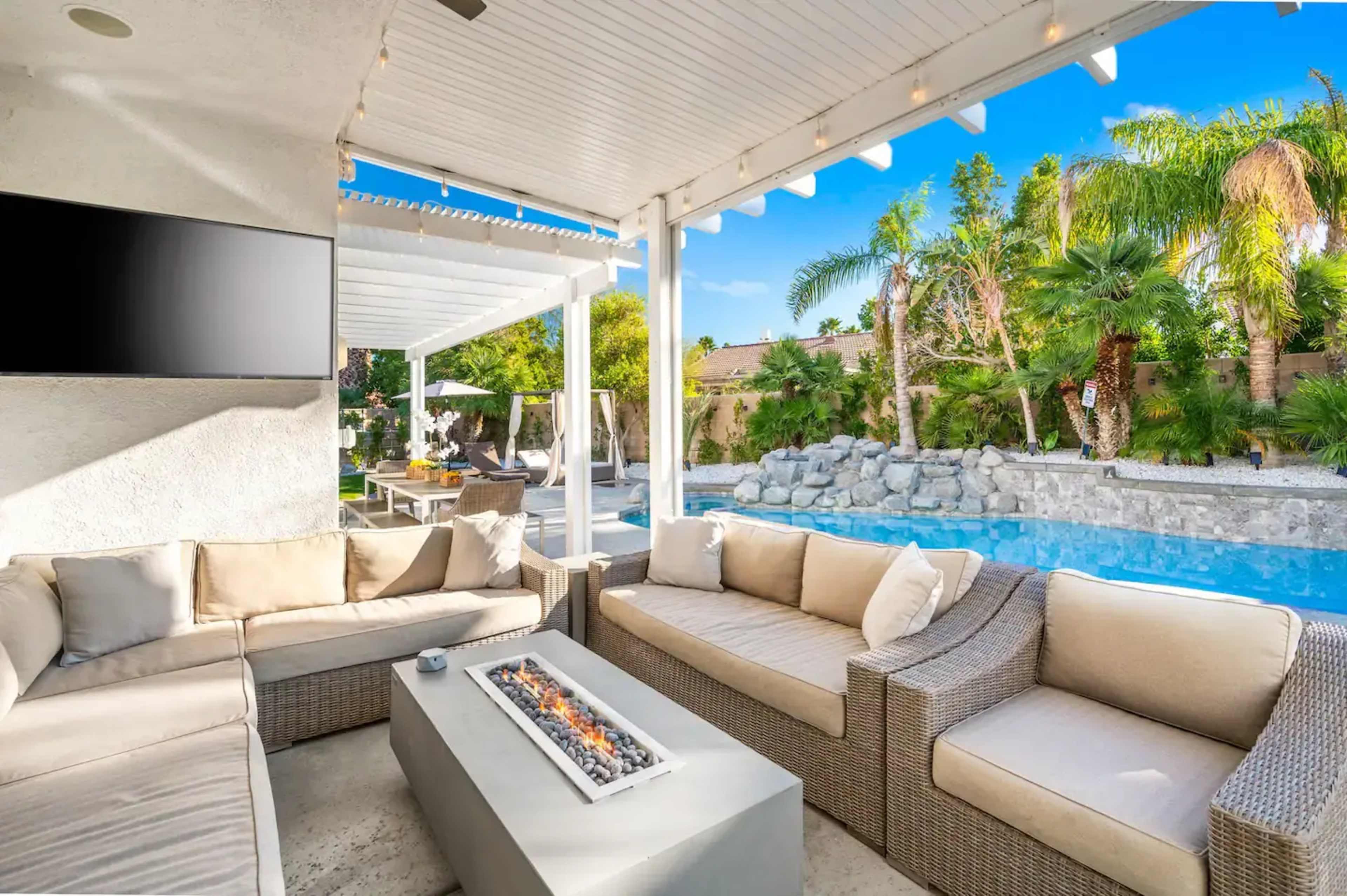 A patio area features a section of modern seating surrounding a fire table, with a swimming pool and palm trees in the background.
