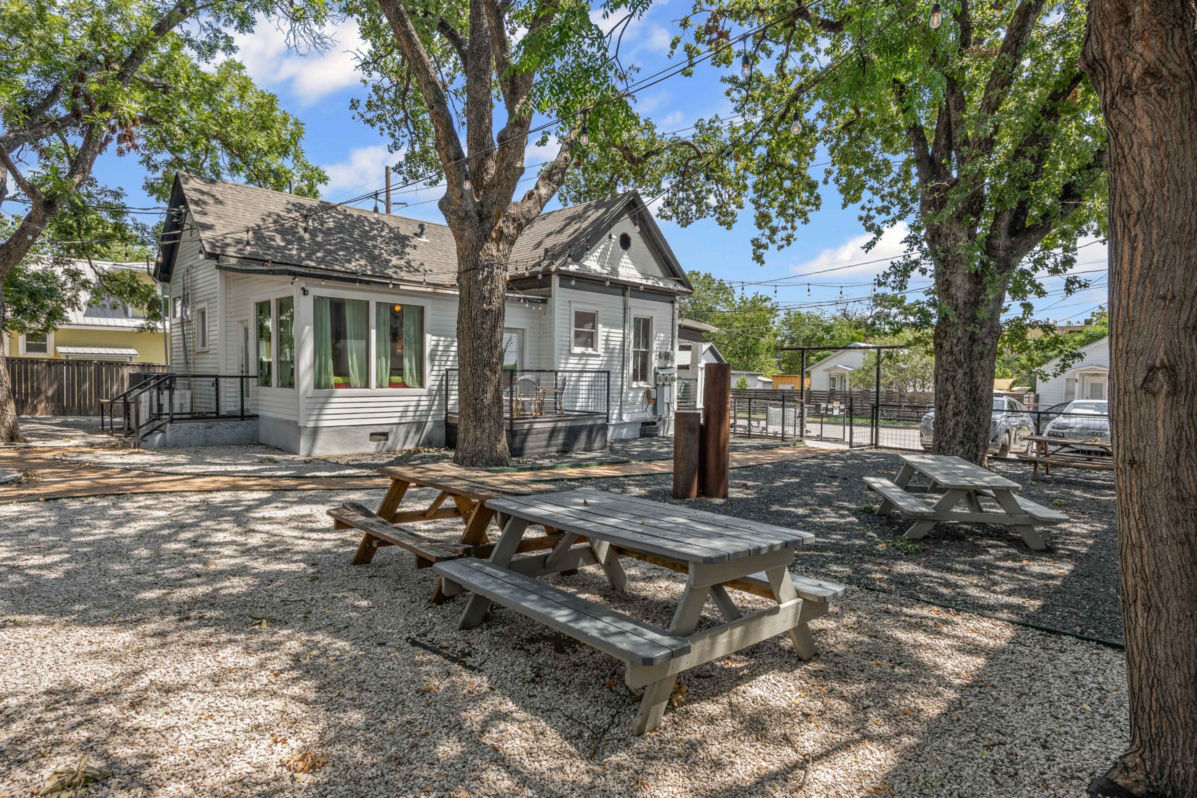 The image shows a light-colored house surrounded by trees, with several picnic tables set on a gravel area in front.