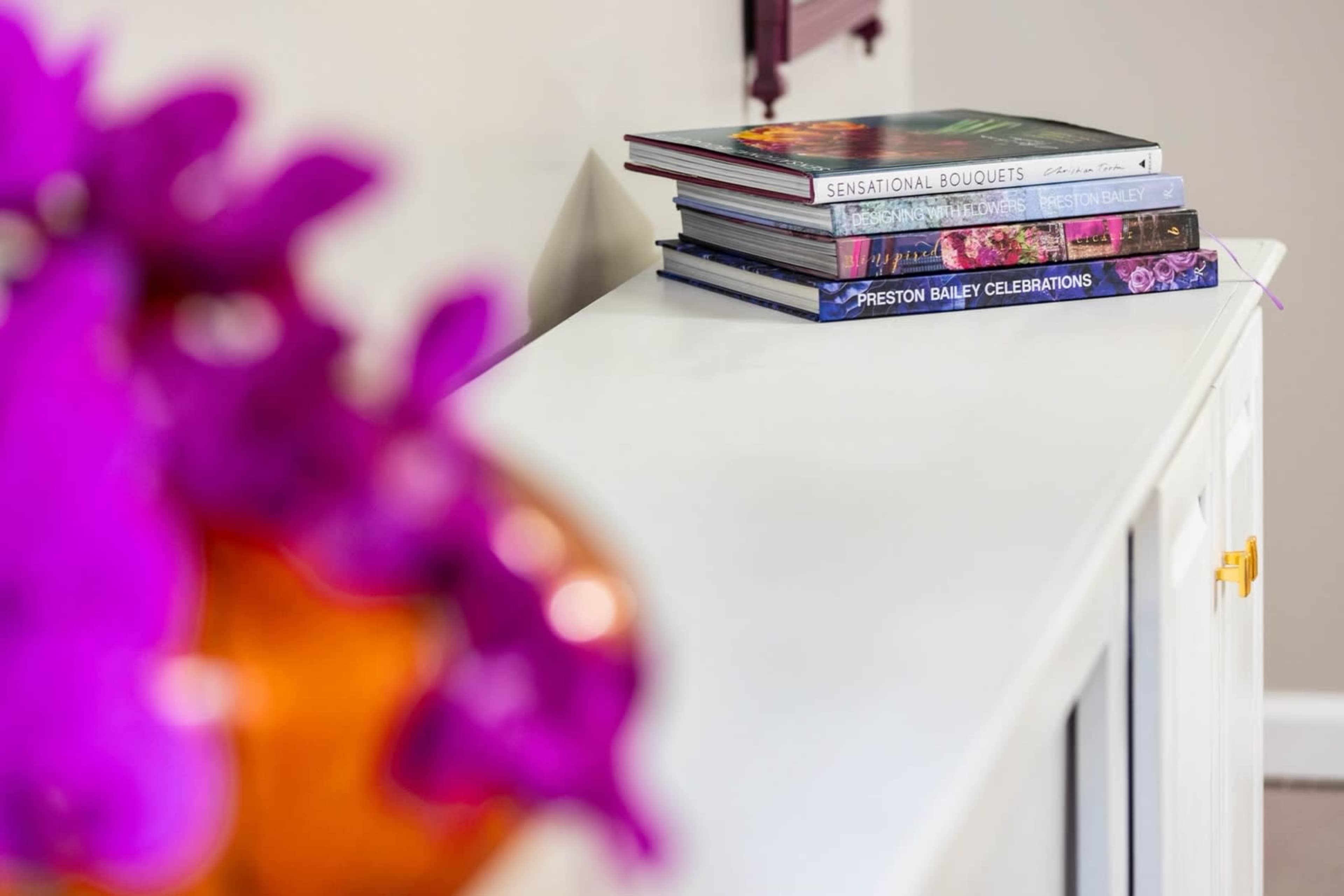 A white console table holds a stack of colorful books and a vase with purple flowers.