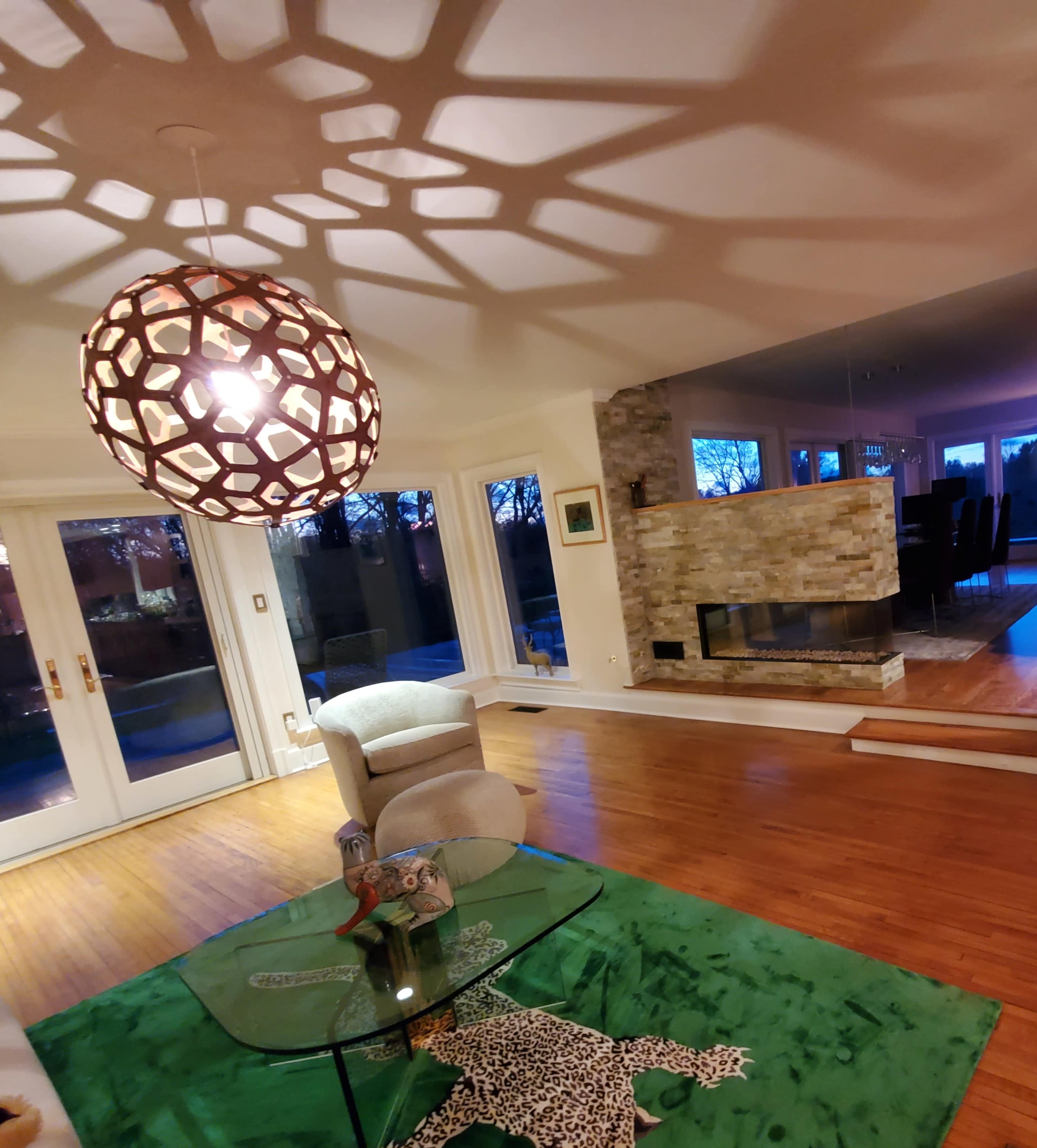 A modern living room with a patterned pendant light casting shadows, a glass coffee table on a green rug, and a stone fireplace visible in the mirrored wall.