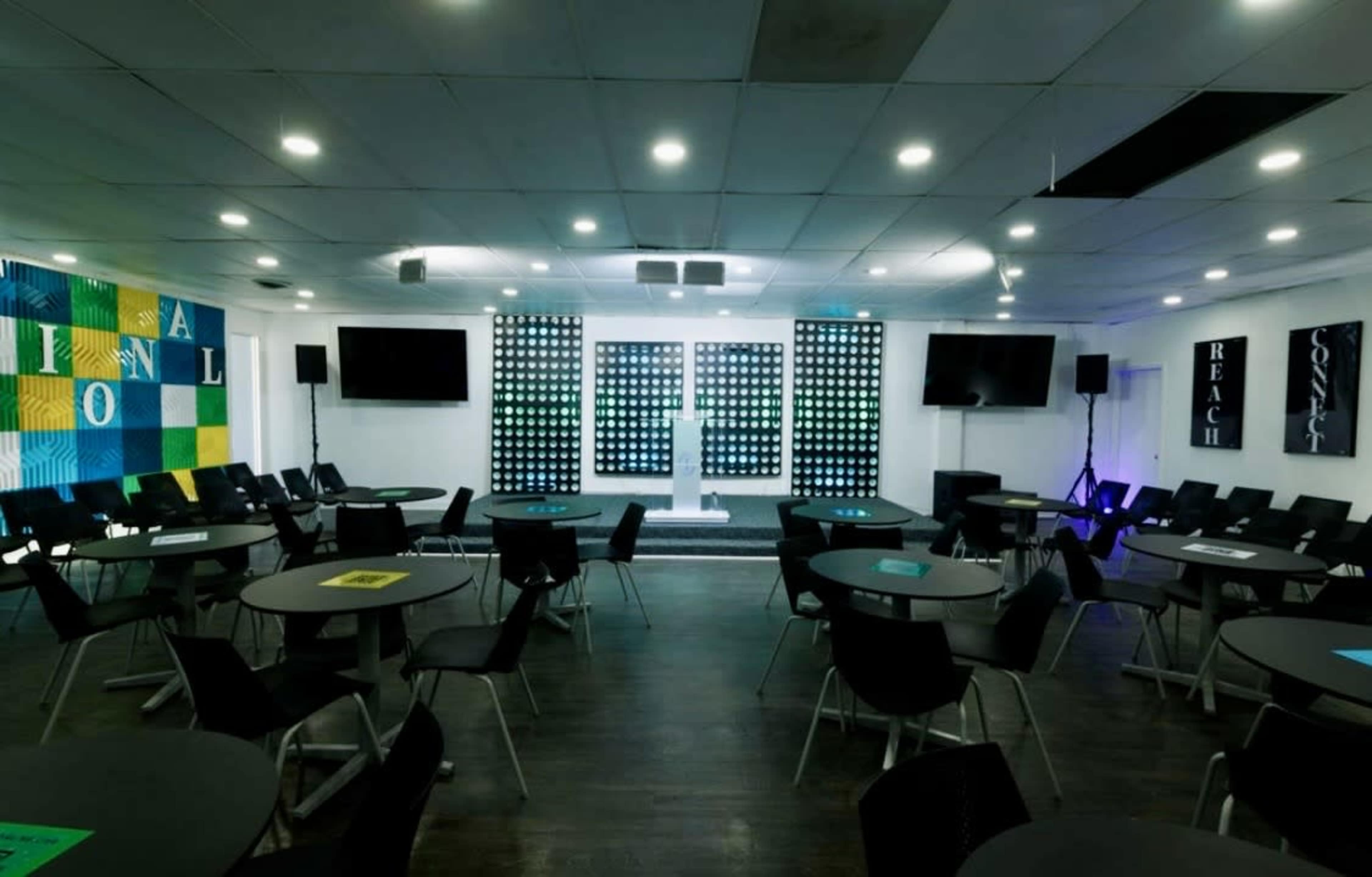 The image shows a spacious event hall with round tables arranged in a seating layout, illuminated by lights and featuring a stage at the front with a backdrop of illuminated panels.