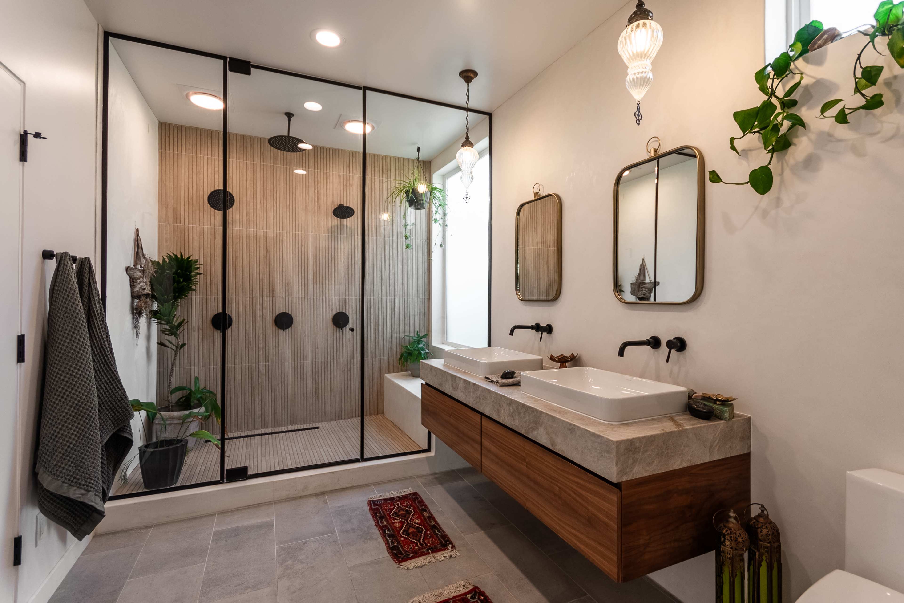 The image shows a modern bathroom with a glass-enclosed shower, dual sink vanity with a wooden base, and decorative plants.