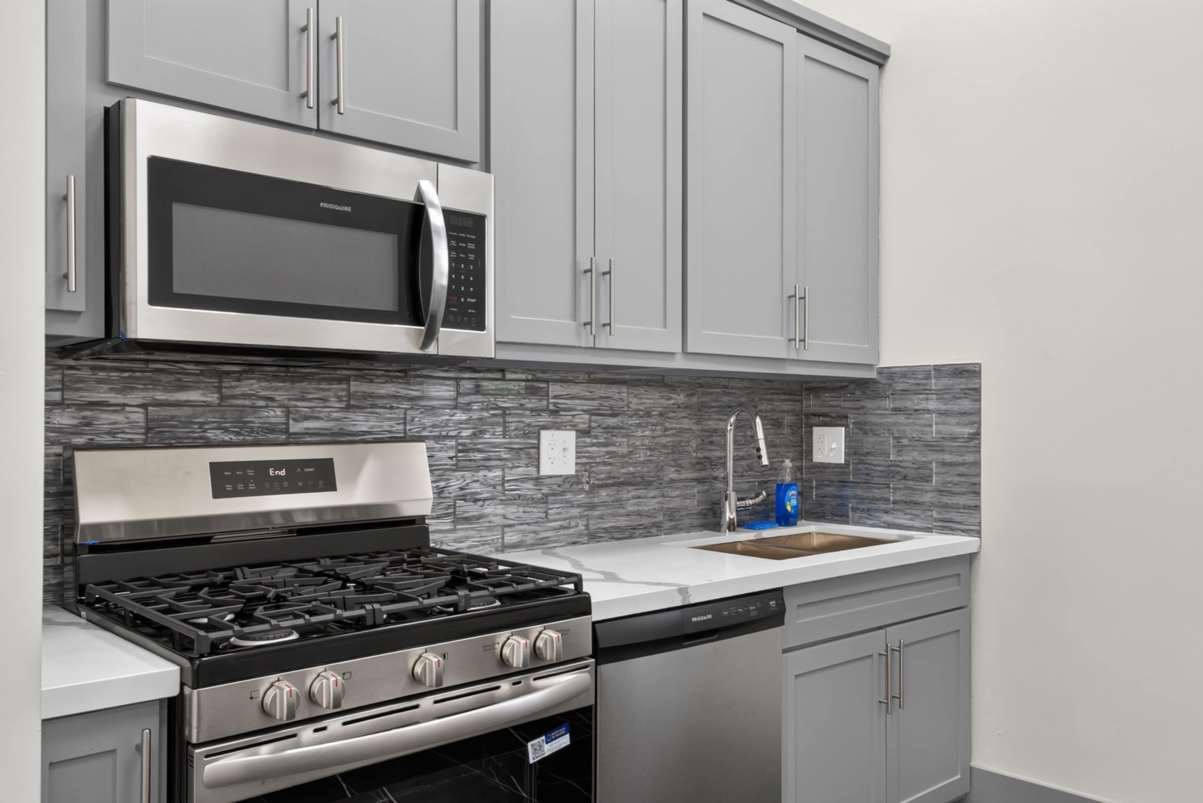 The image shows a modern kitchen featuring stainless steel appliances, including a microwave and a gas stove, with gray cabinetry and a stone-patterned backsplash.