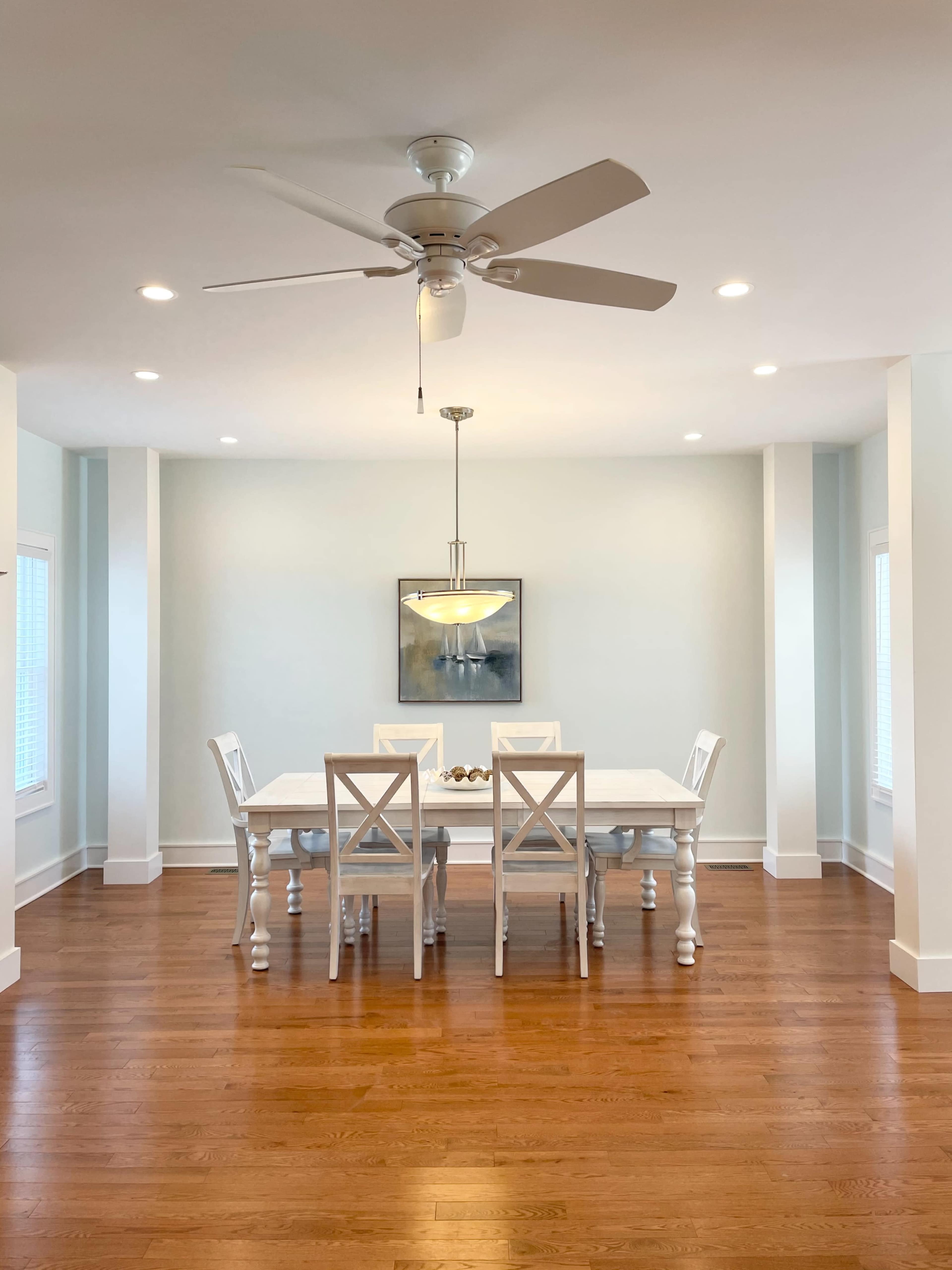A bright dining room features a white table with six chairs, a ceiling fan above, and light hardwood floors.