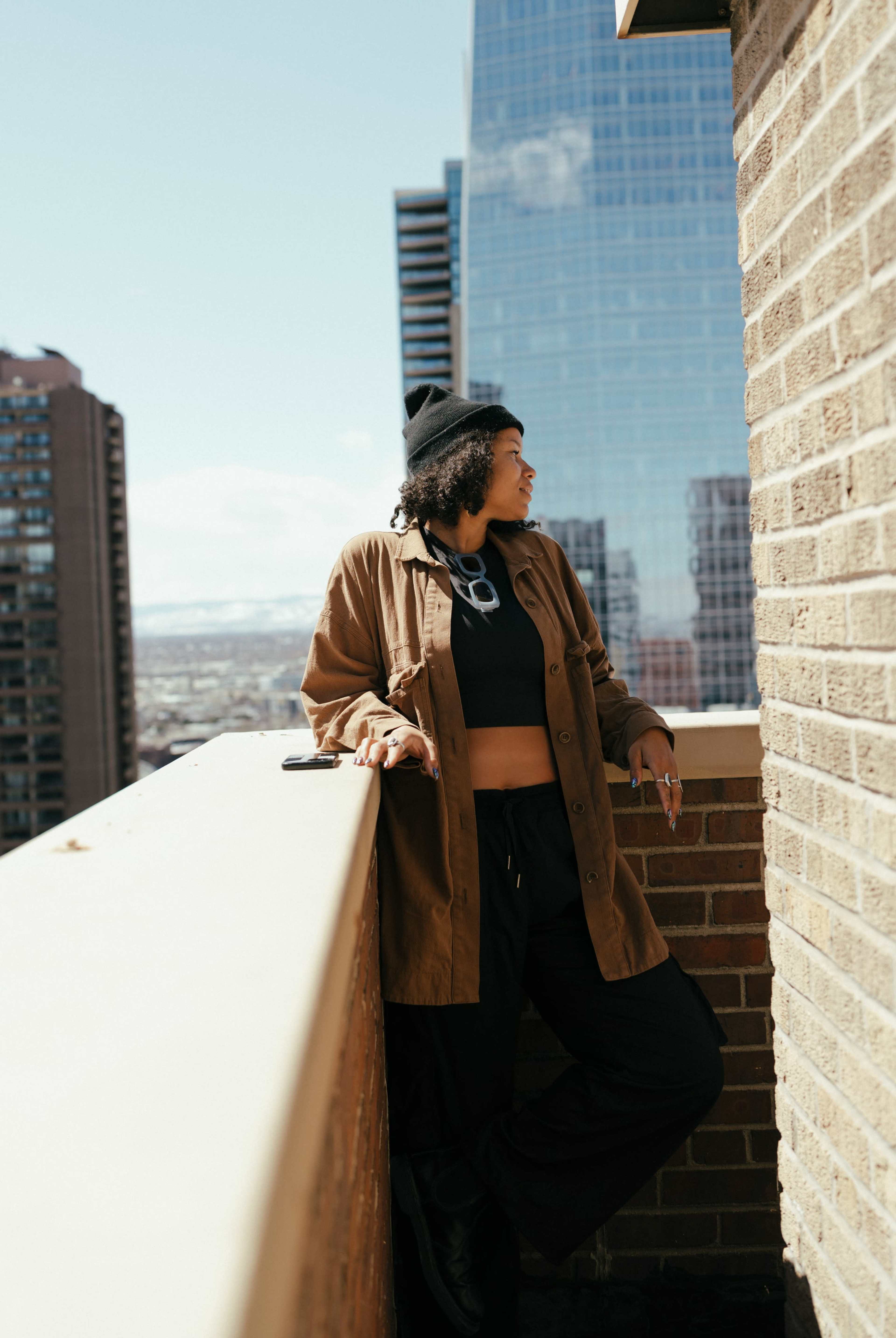A woman stands casually on a balcony, overlooking a cityscape, wearing a brown jacket and black pants.