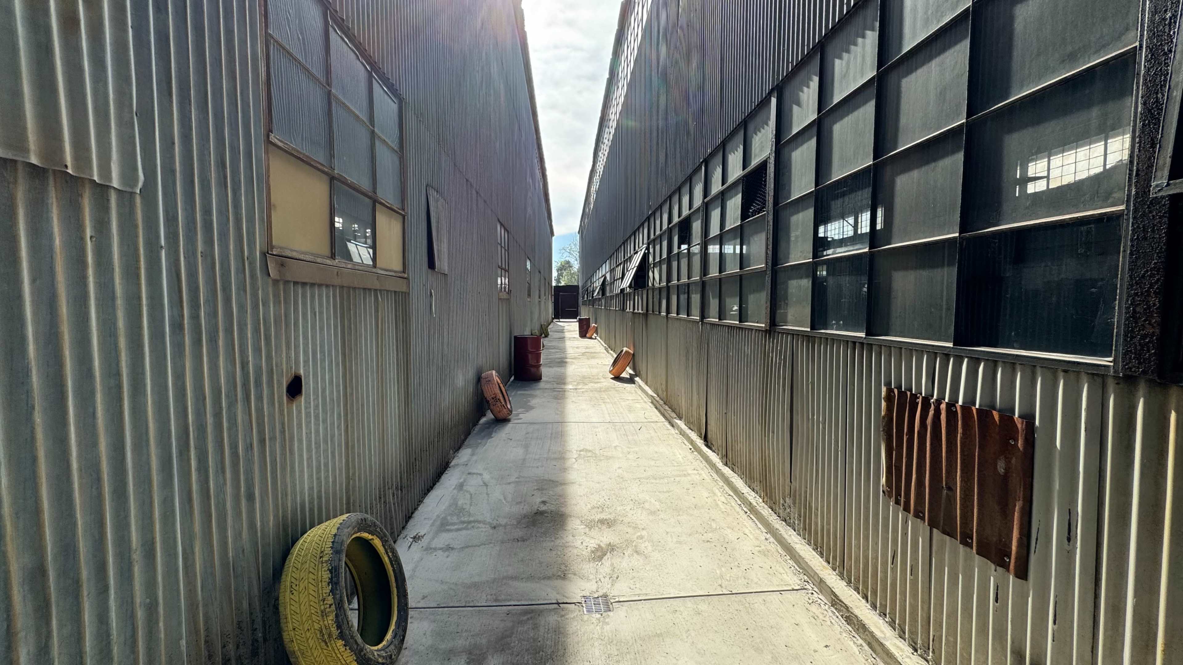 A narrow alleyway lined with corrugated metal buildings and large windows leads to a brightly lit end.