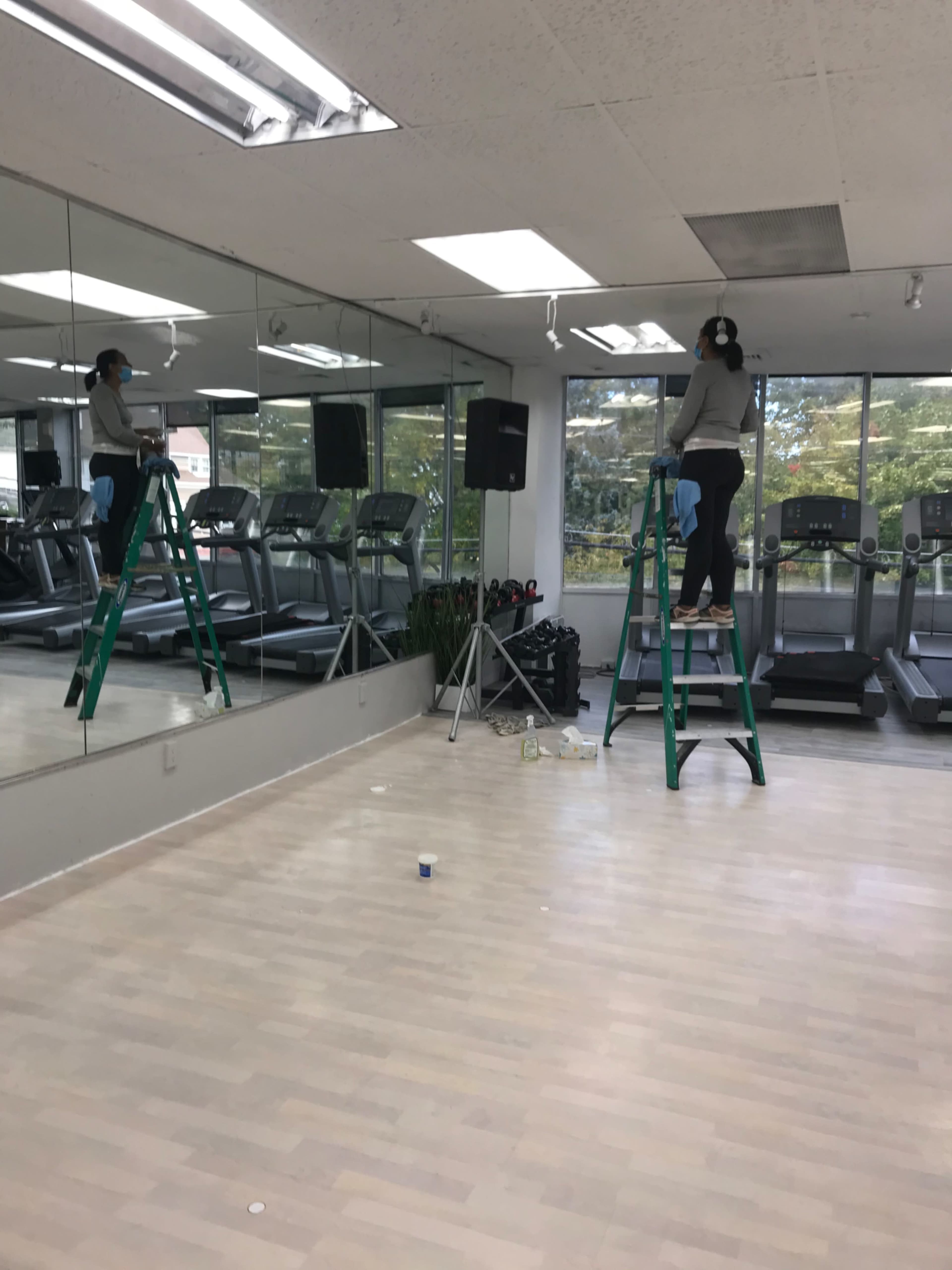 A person stands on a green ladder installing equipment in a mirror-lined gym with treadmills visible in the background.