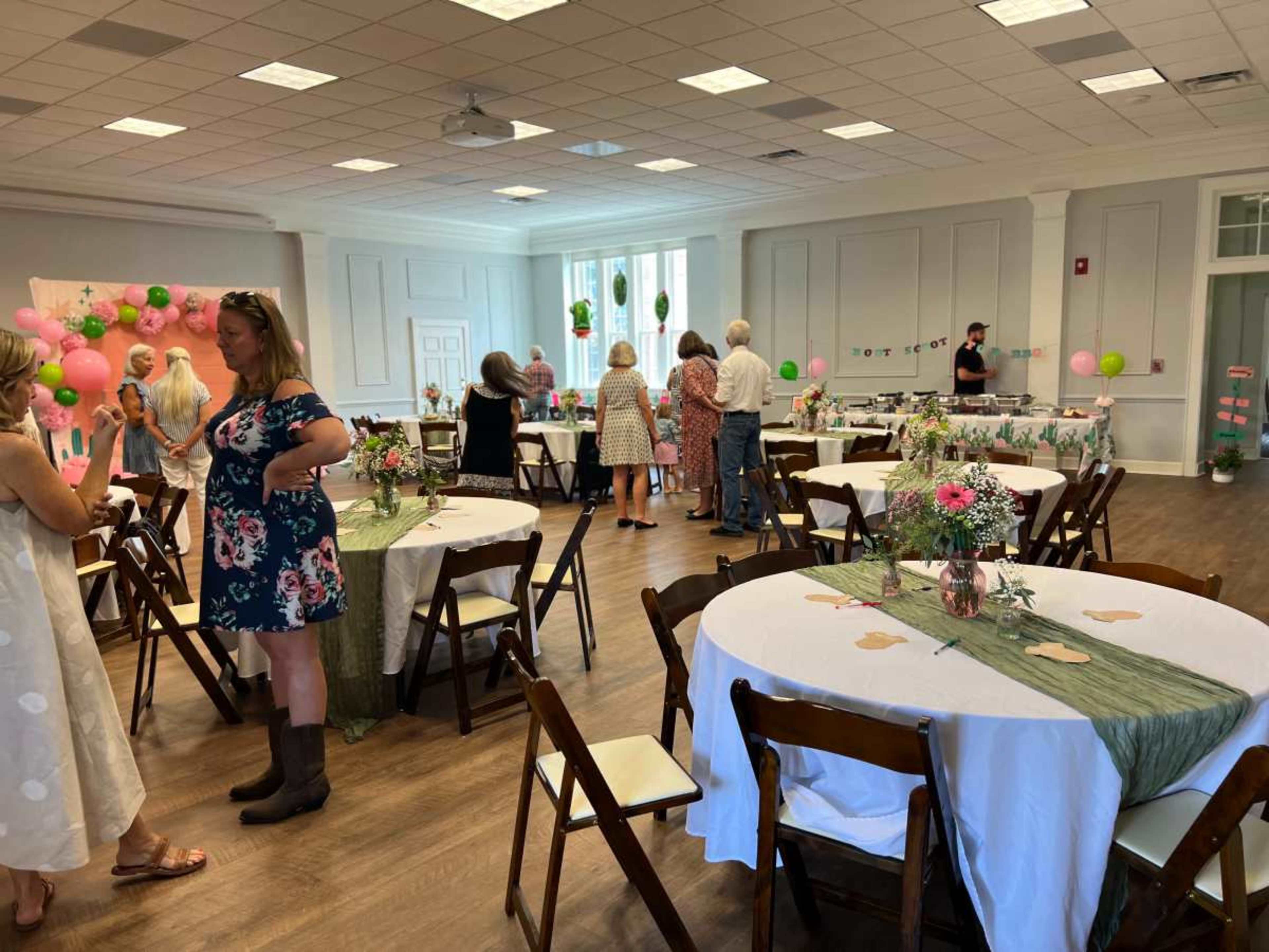 A bright event space with round tables covered in white and green cloths, decorated with flowers, while guests socialize and partake in a gathering.
