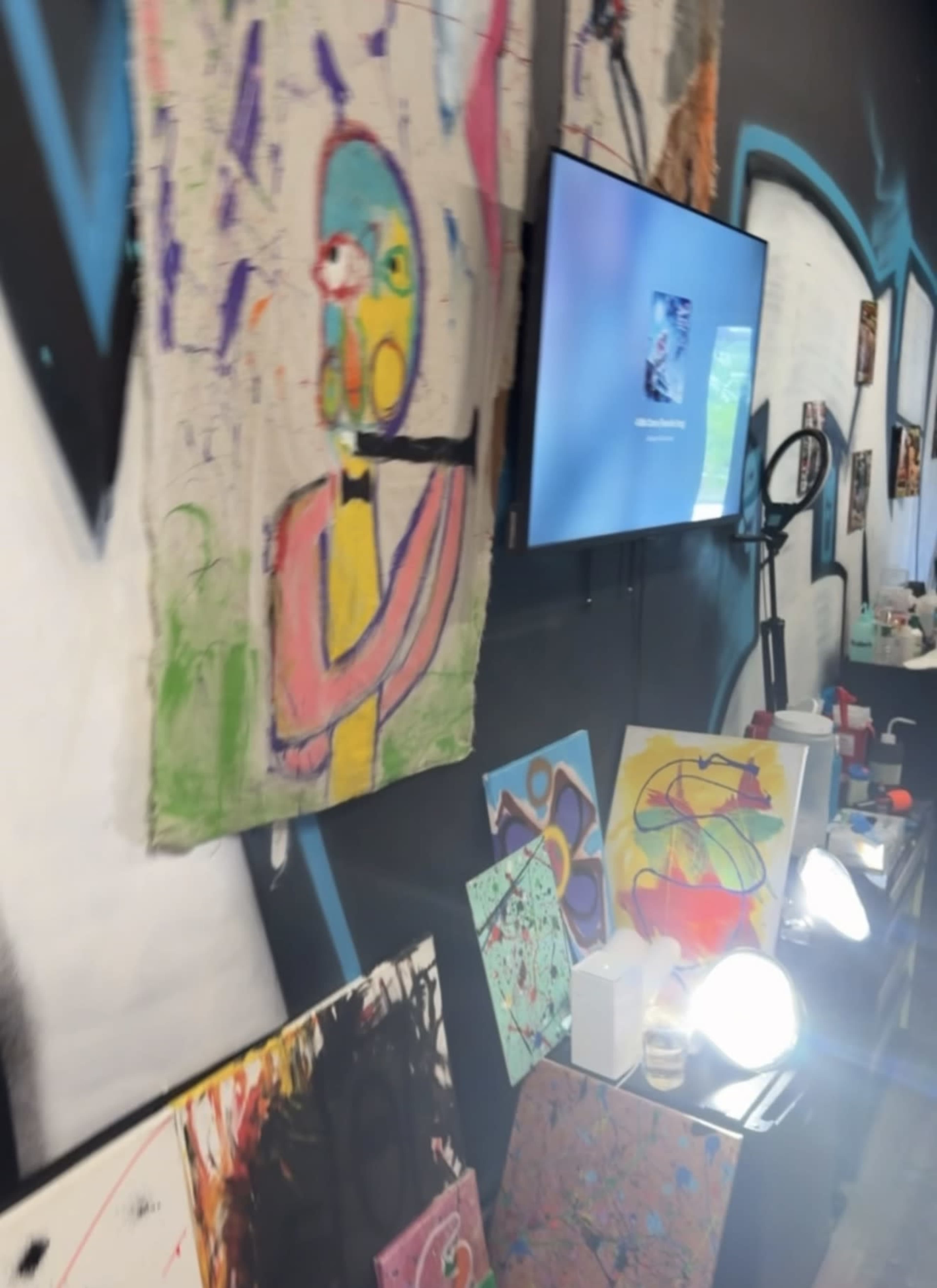 The image shows a colorful wall adorned with abstract paintings and a television set, alongside a display of various art pieces on a table below.