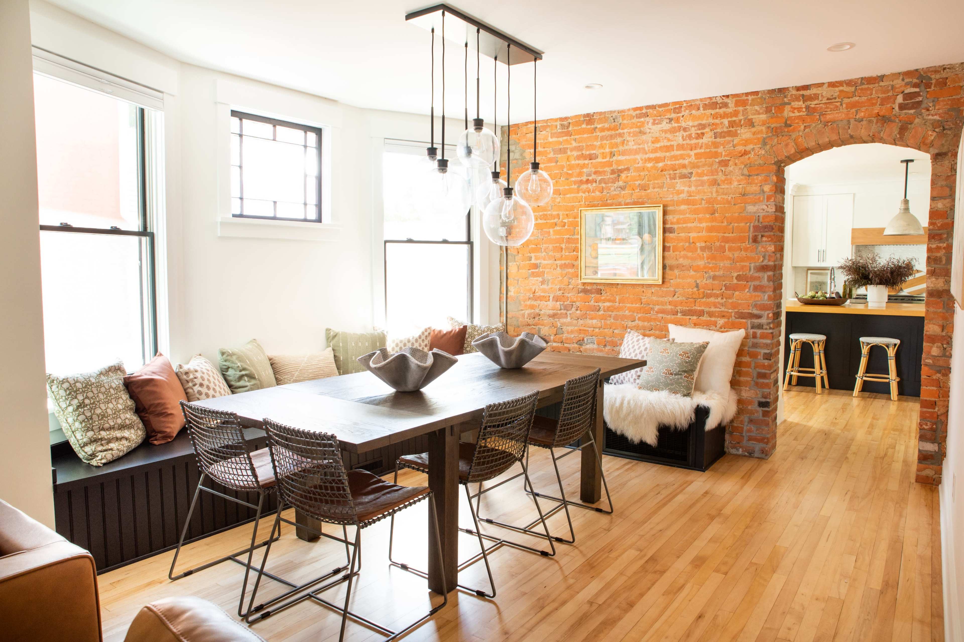 A dining area with a wooden table surrounded by wire chairs, a brick wall, and large windows illuminating the space.