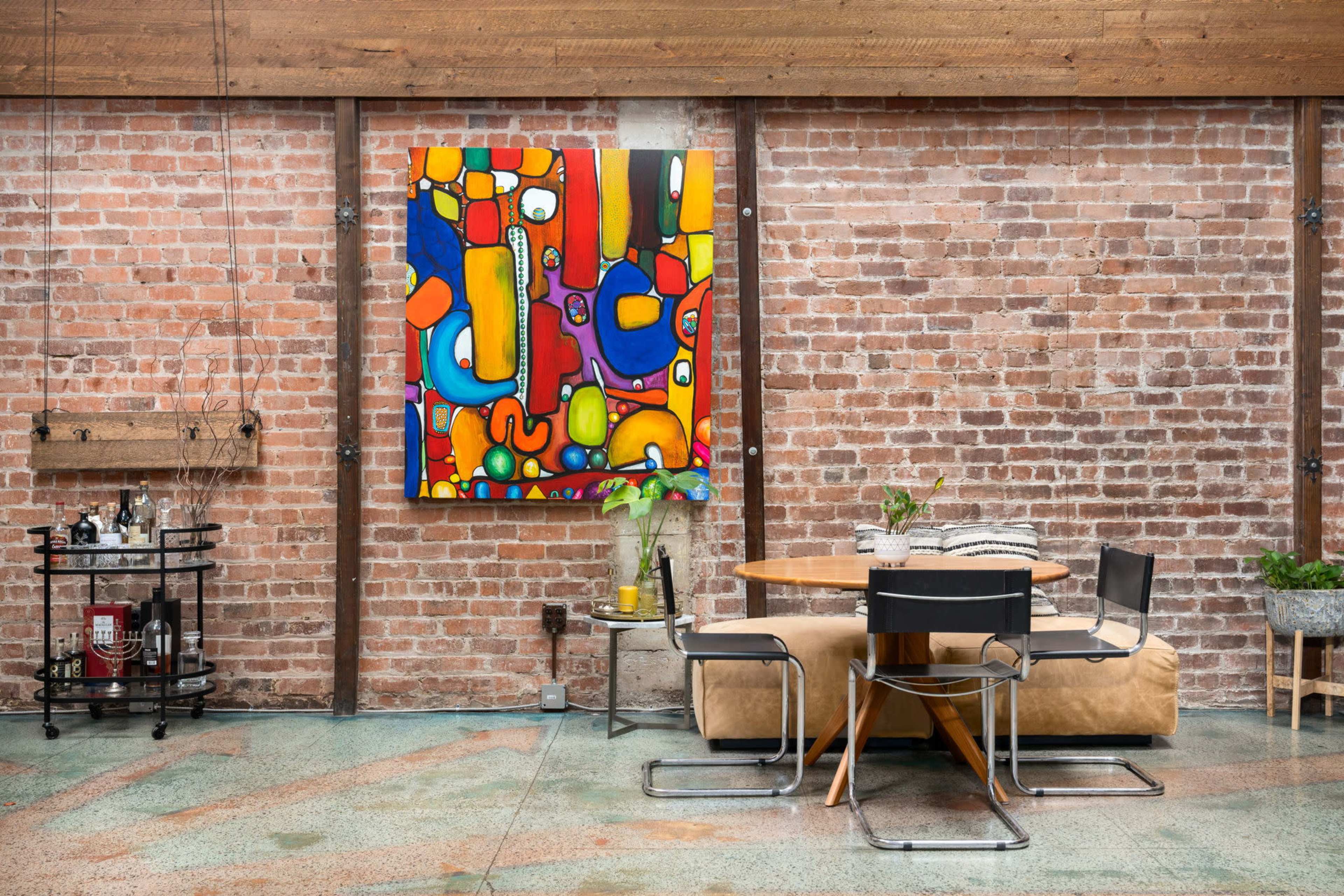 A round wooden dining table with black chairs is positioned against a brick wall featuring a colorful abstract painting.