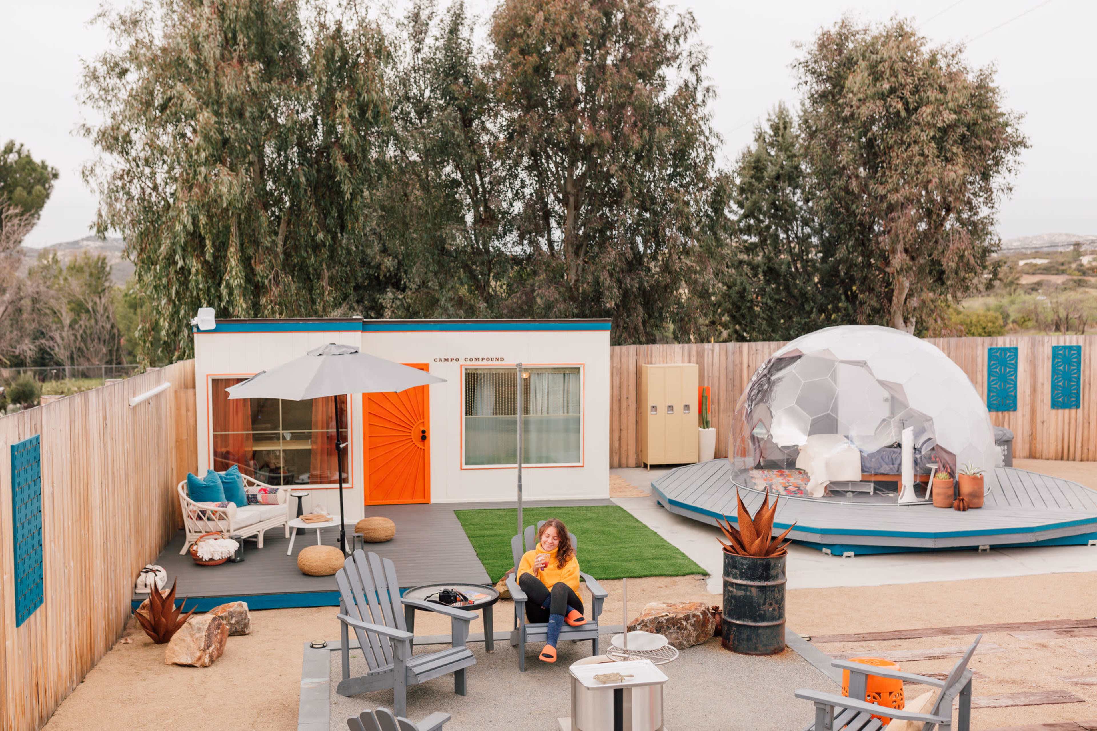 A person sits on a chair in a backyard area with a modern container-style home and a geodesic dome structure nearby.