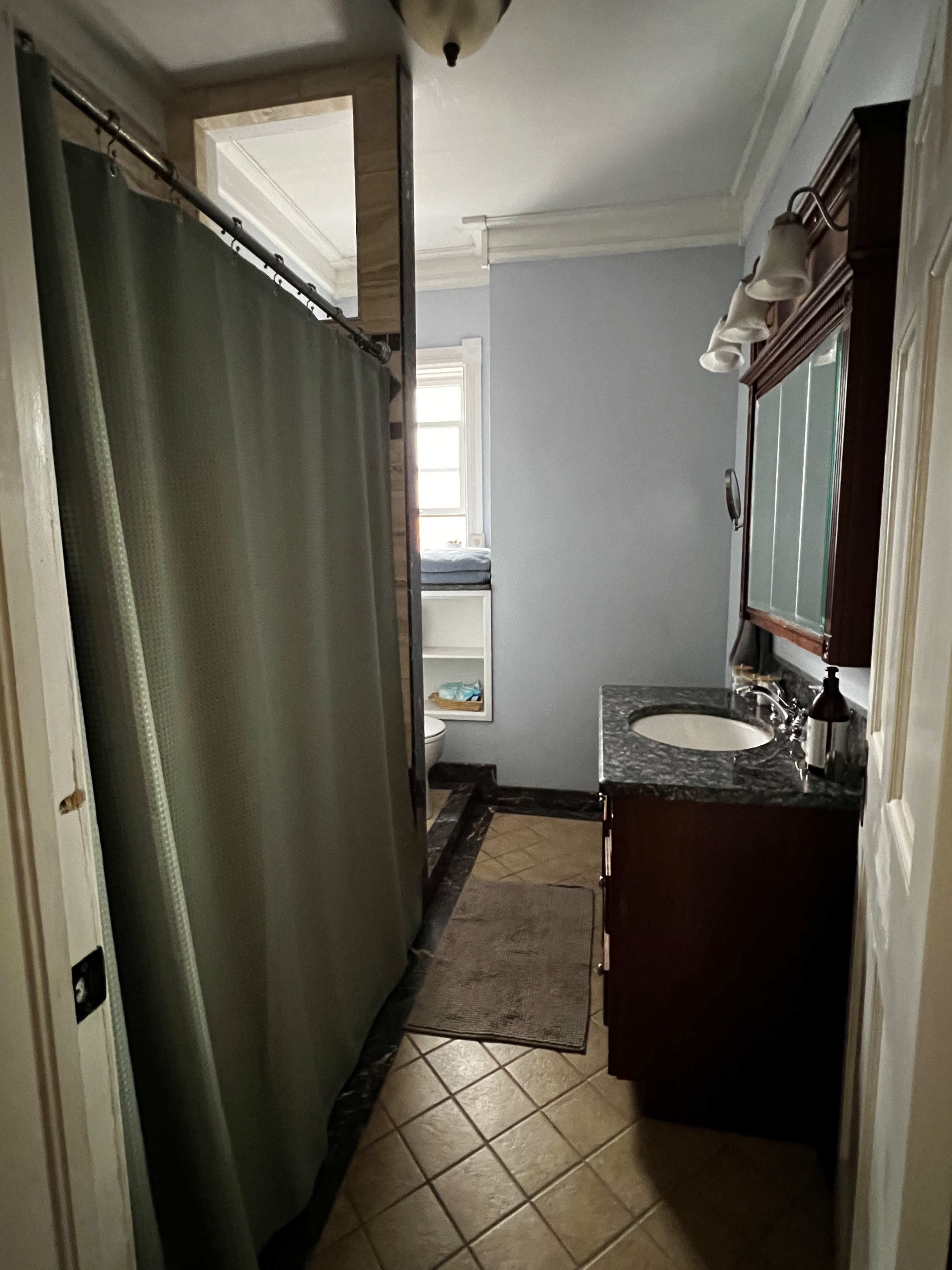 The image shows a bathroom featuring a shower curtain, a sink with a mirror above it, and a toilet positioned near a window.