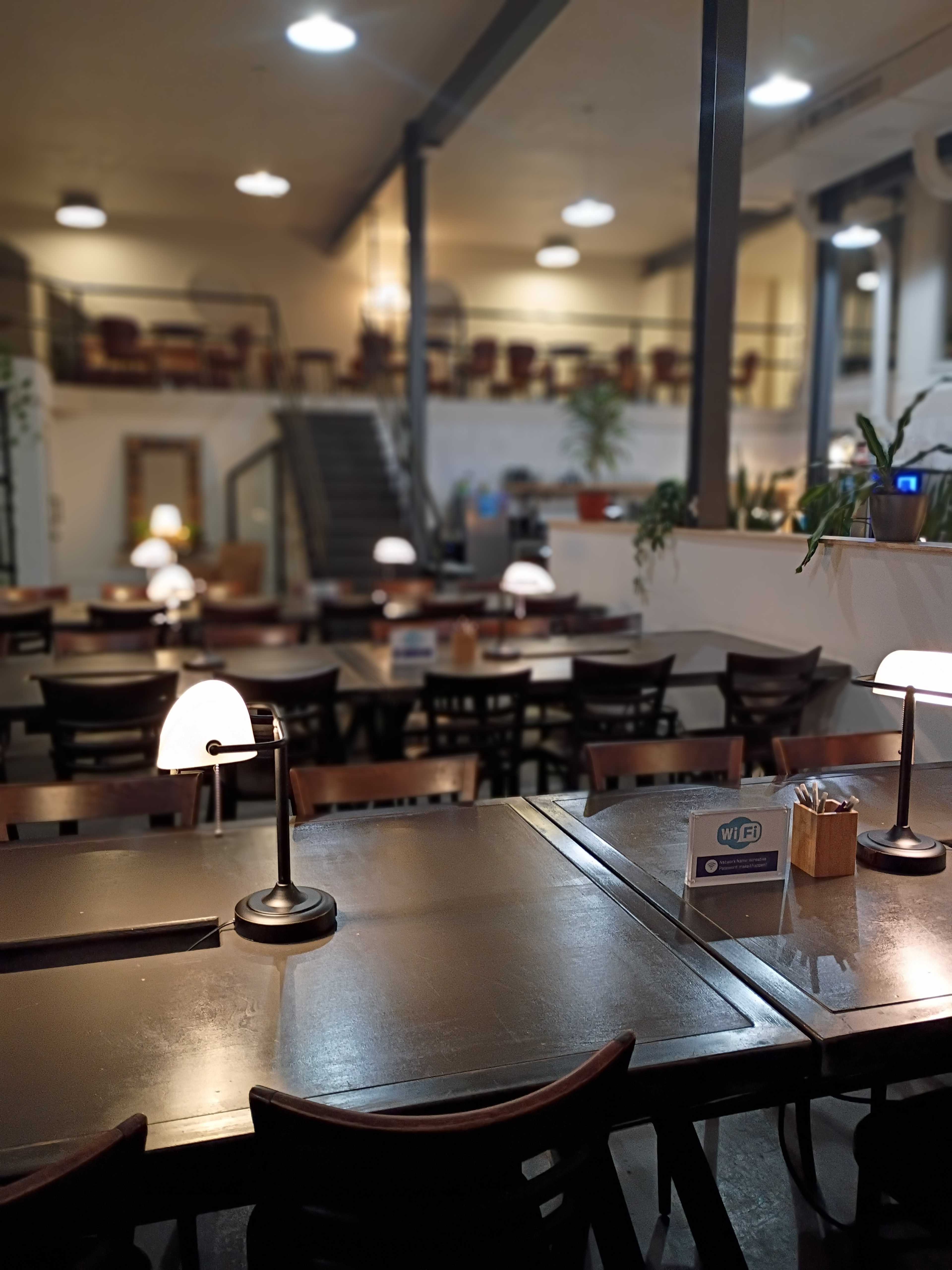 The image shows an interior of a restaurant with multiple tables, a staircase in the background, and lamps on each table providing ambient lighting.