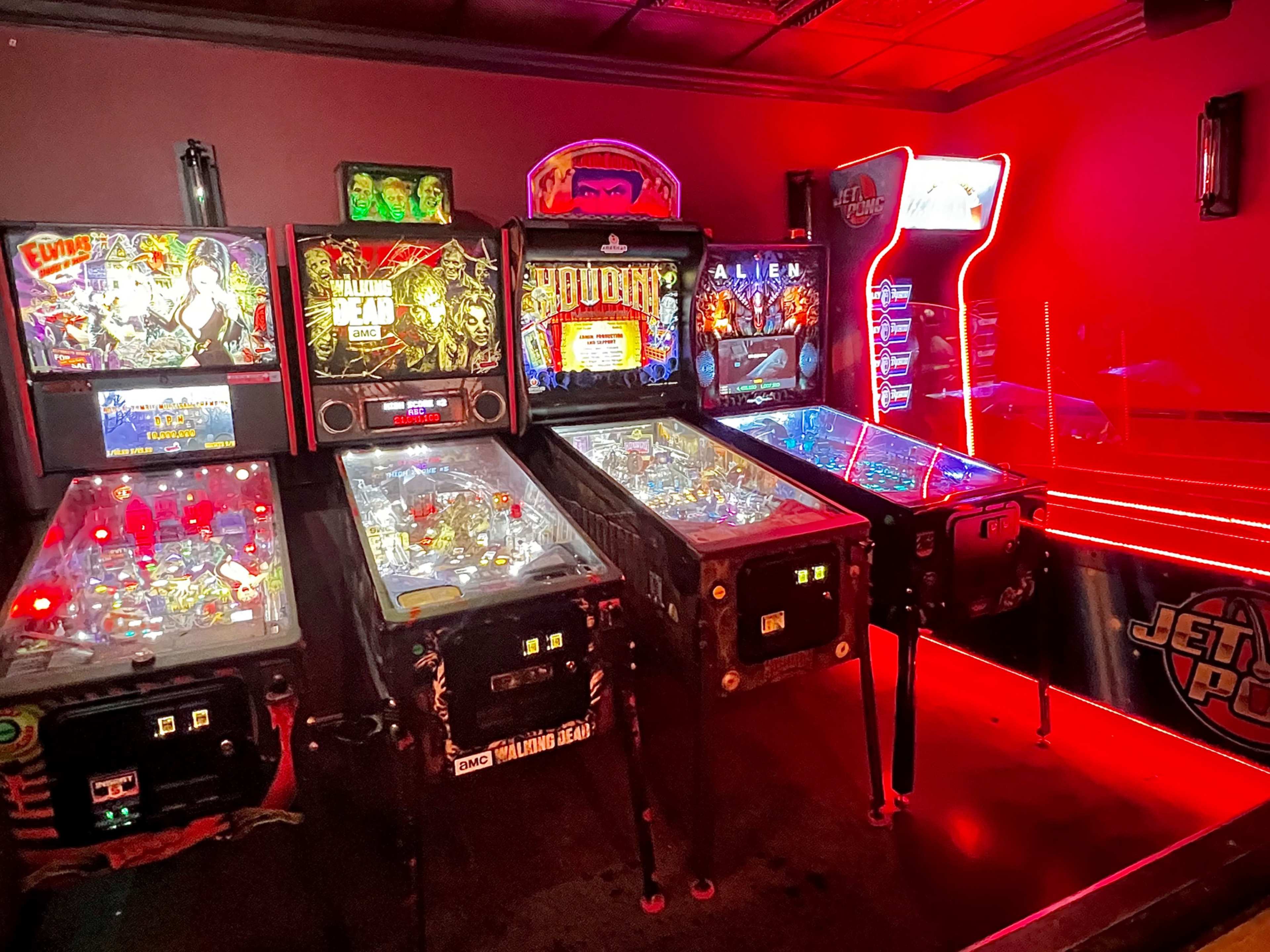 The image shows a lineup of pinball machines illuminated by colorful lights in a dimly lit arcade.