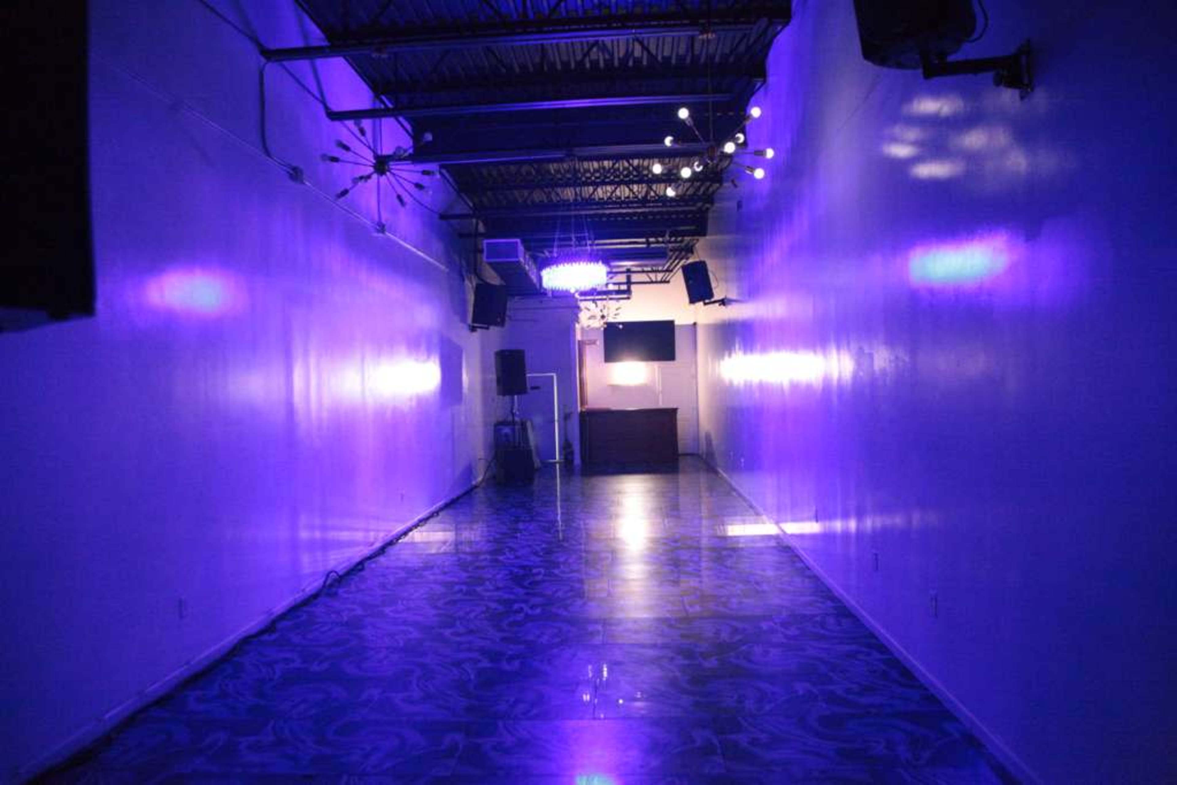 The image shows a long, narrow room with purple lighting, a polished floor, and a DJ booth at the far end.
