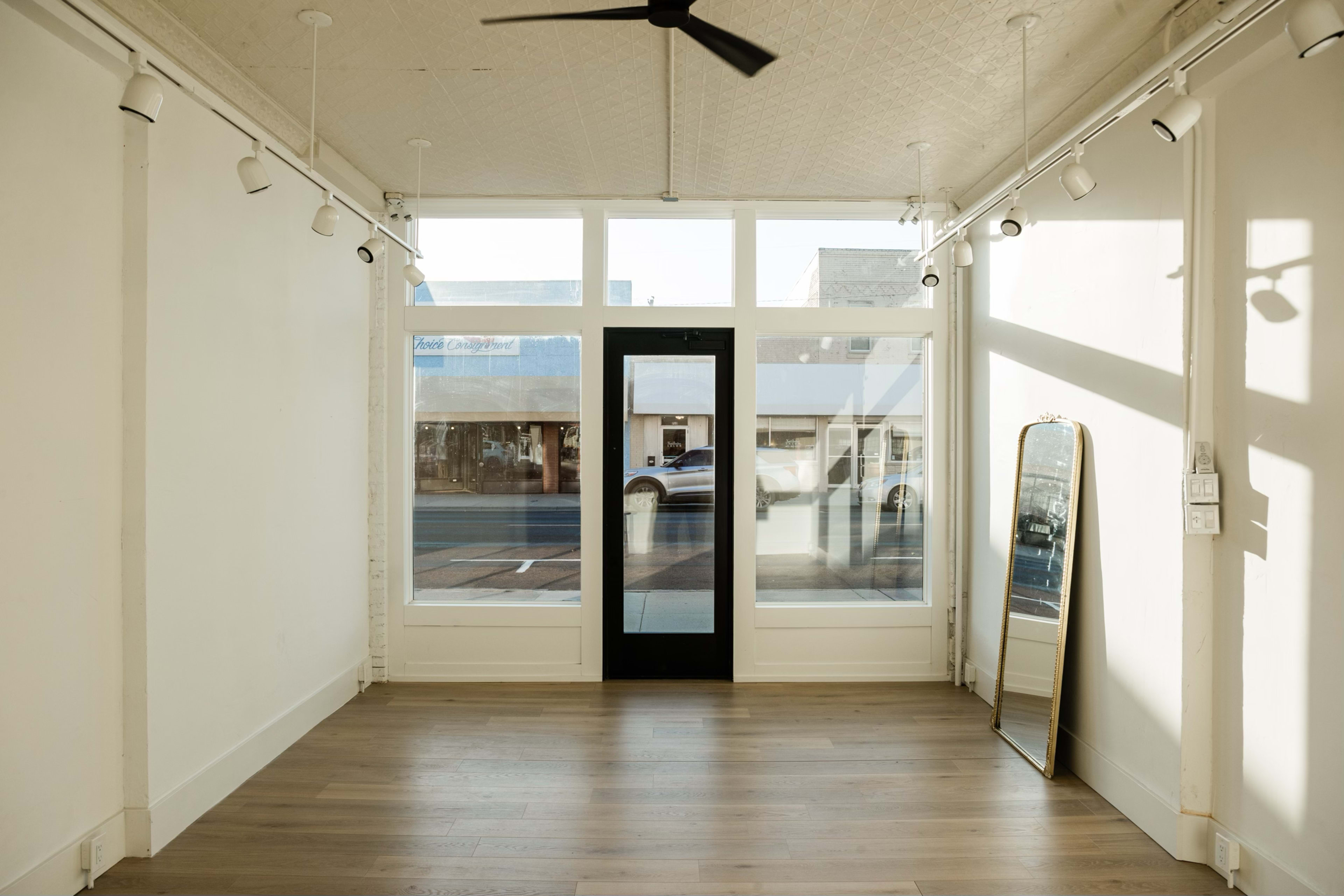 The image shows an empty room with large windows, a black door, and a tall mirror against the wall.