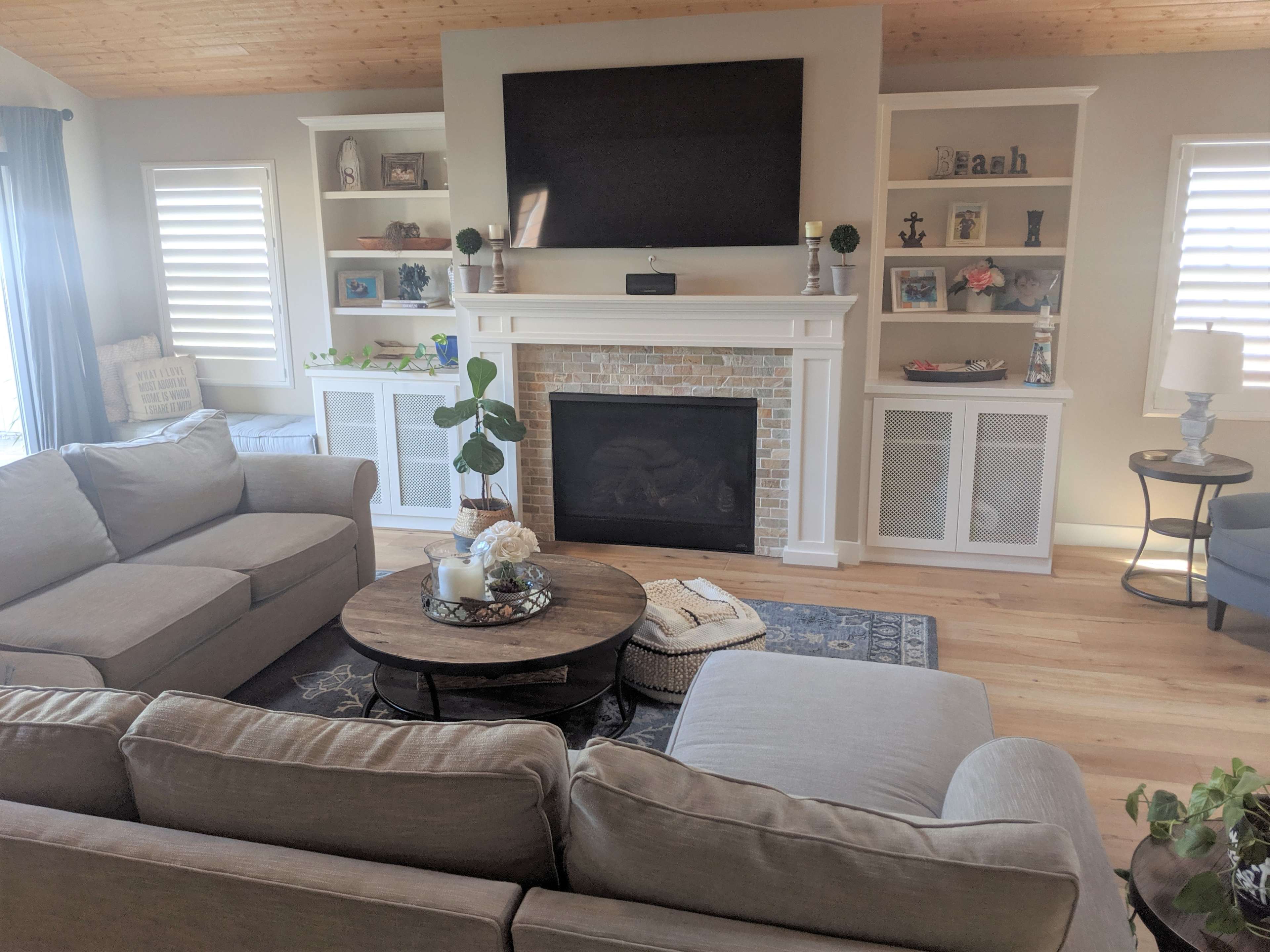 The image shows a living room with two gray sofas facing a circular coffee table, a stone fireplace beneath a mounted television, and built-in shelves displaying various items.