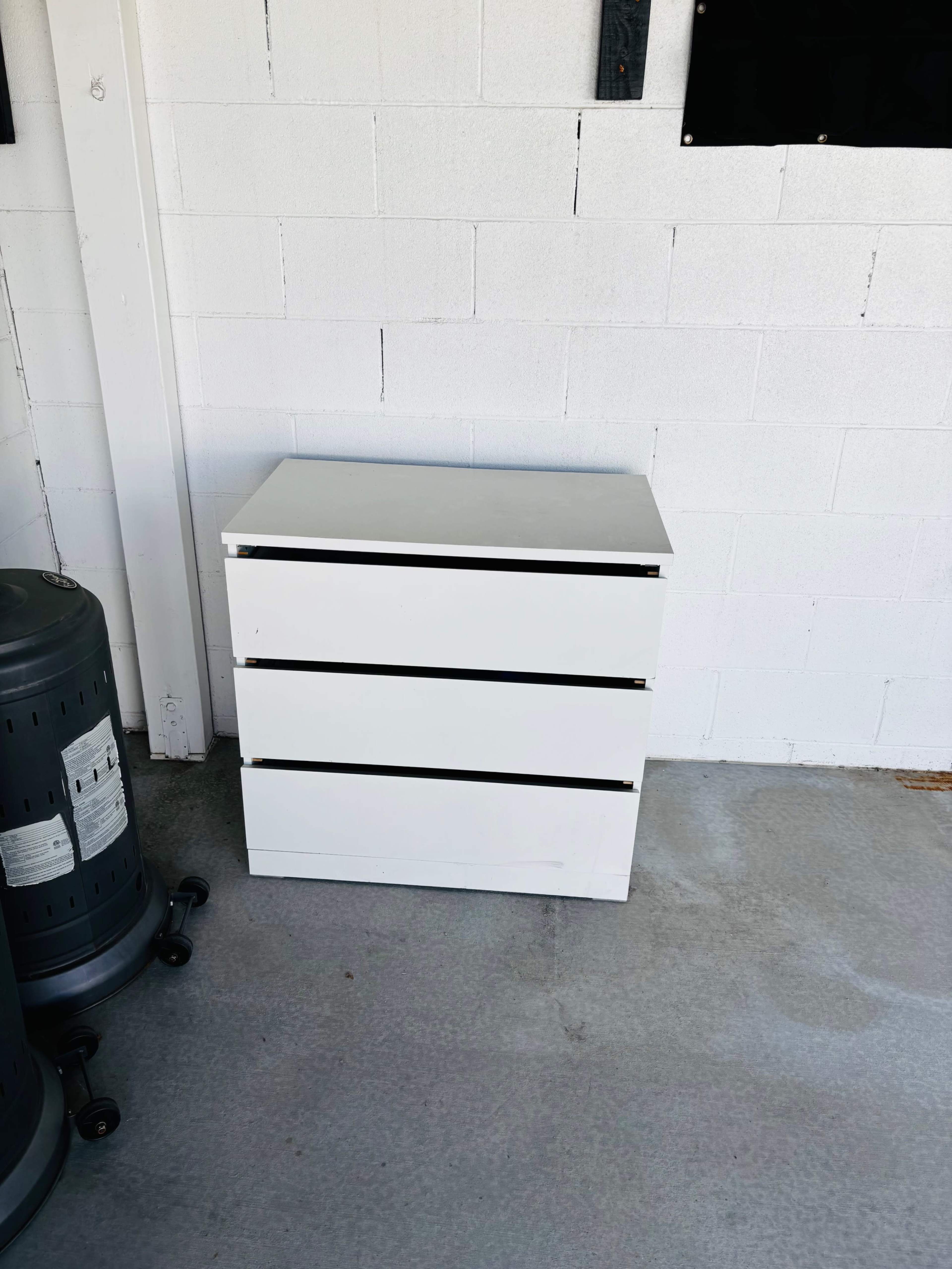 A white three-drawer chest is positioned against a white cinder block wall next to a black cylindrical object.