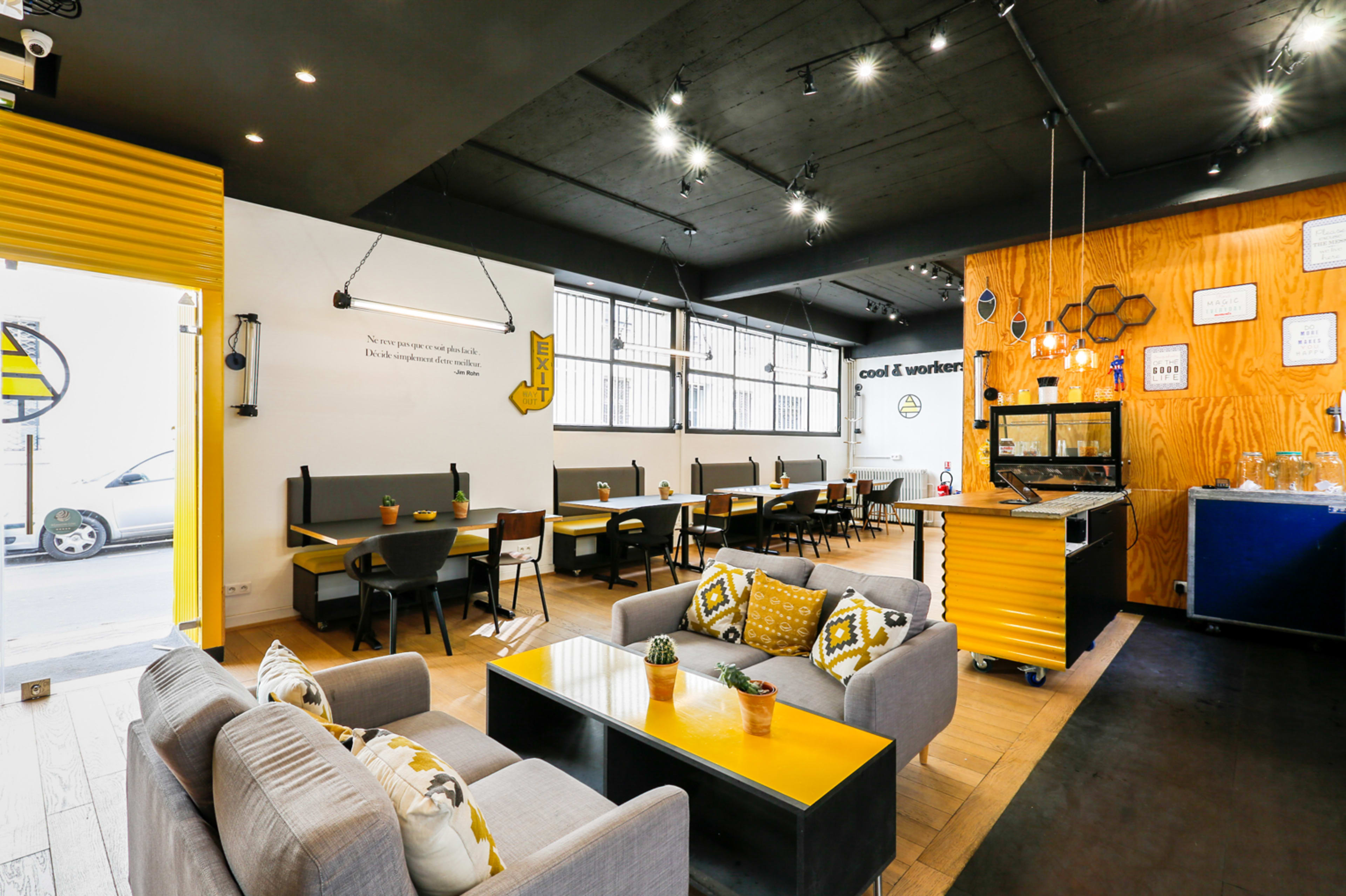The image shows a modern café with wooden seating, tables, potted plants, and yellow accents, illuminated by overhead lights.