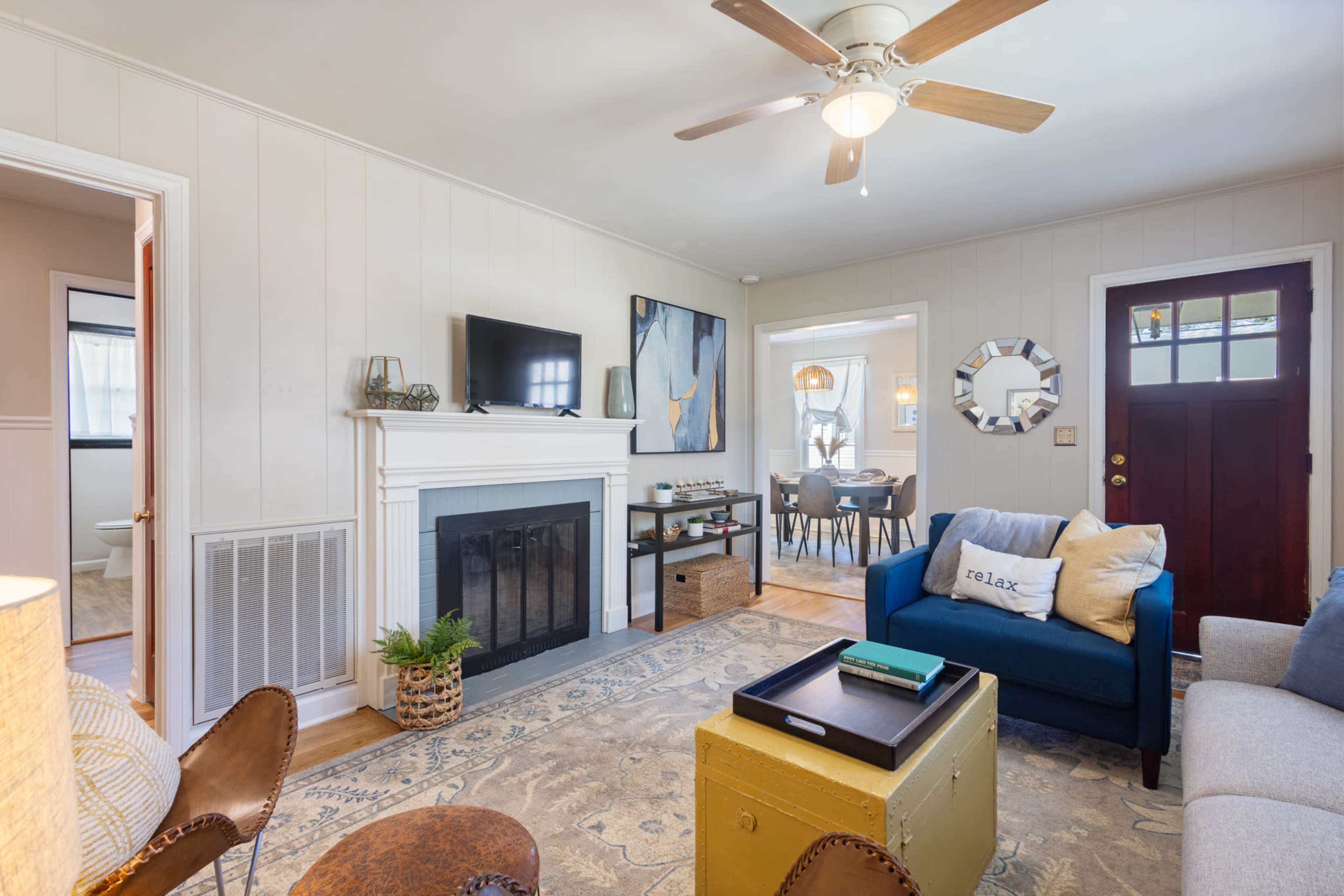 A cozy living room features a blue sofa, a coffee table, and a fireplace with a television mounted above it, leading to a dining area through an open doorway.