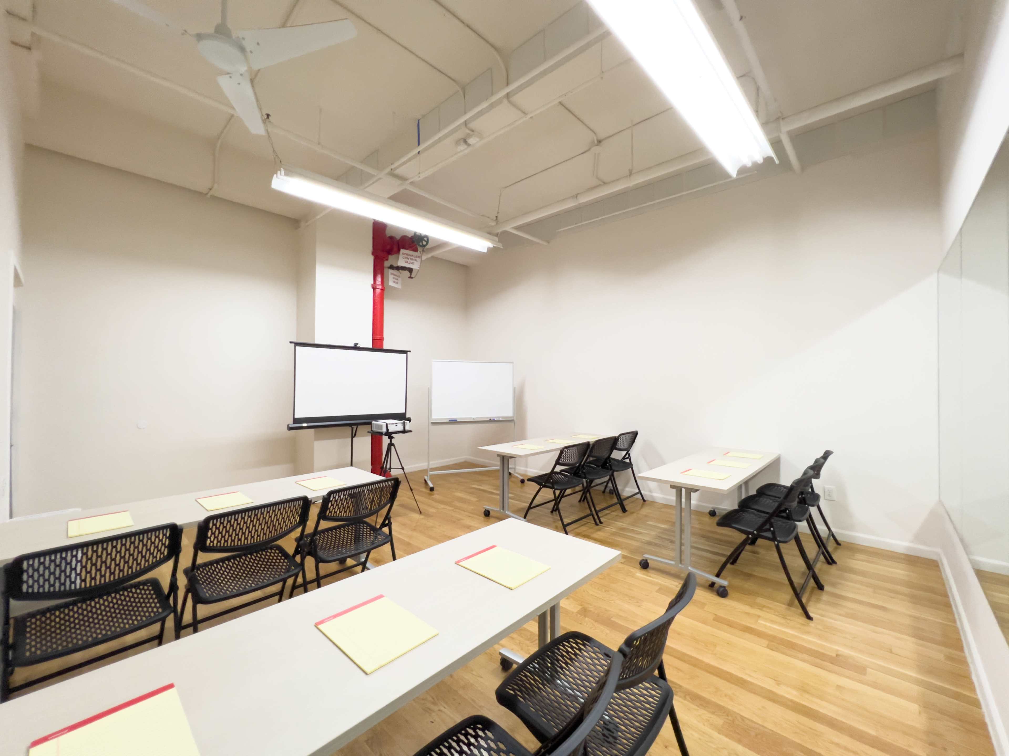 A spacious meeting room with several tables and chairs arranged around two whiteboards and a projector screen.