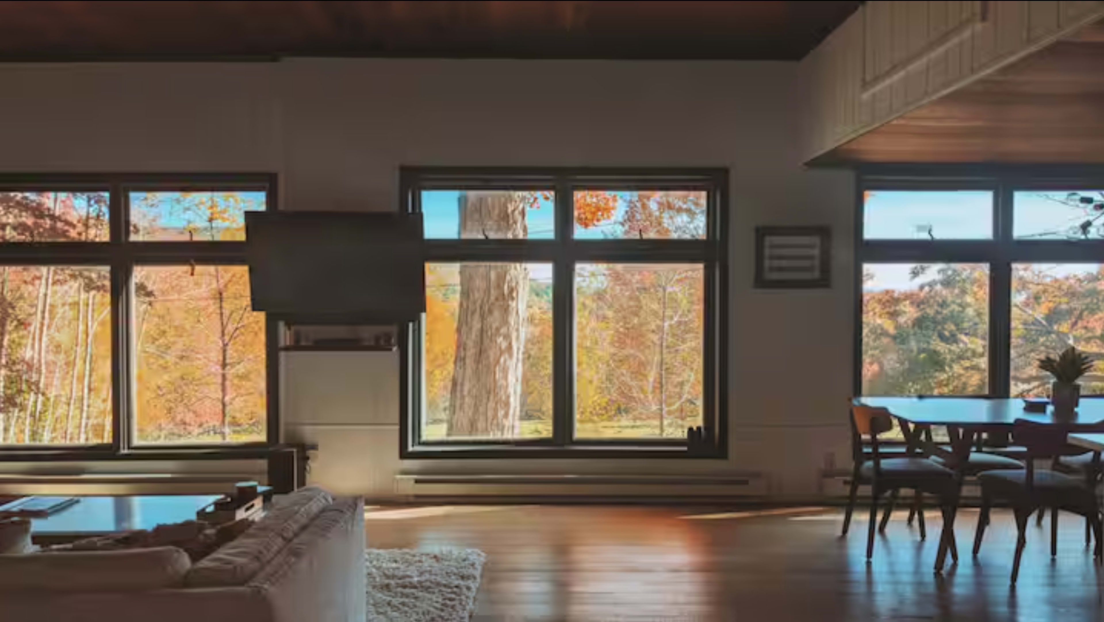 The image shows a modern living room with large windows framing a view of colorful autumn foliage and a cozy seating area.