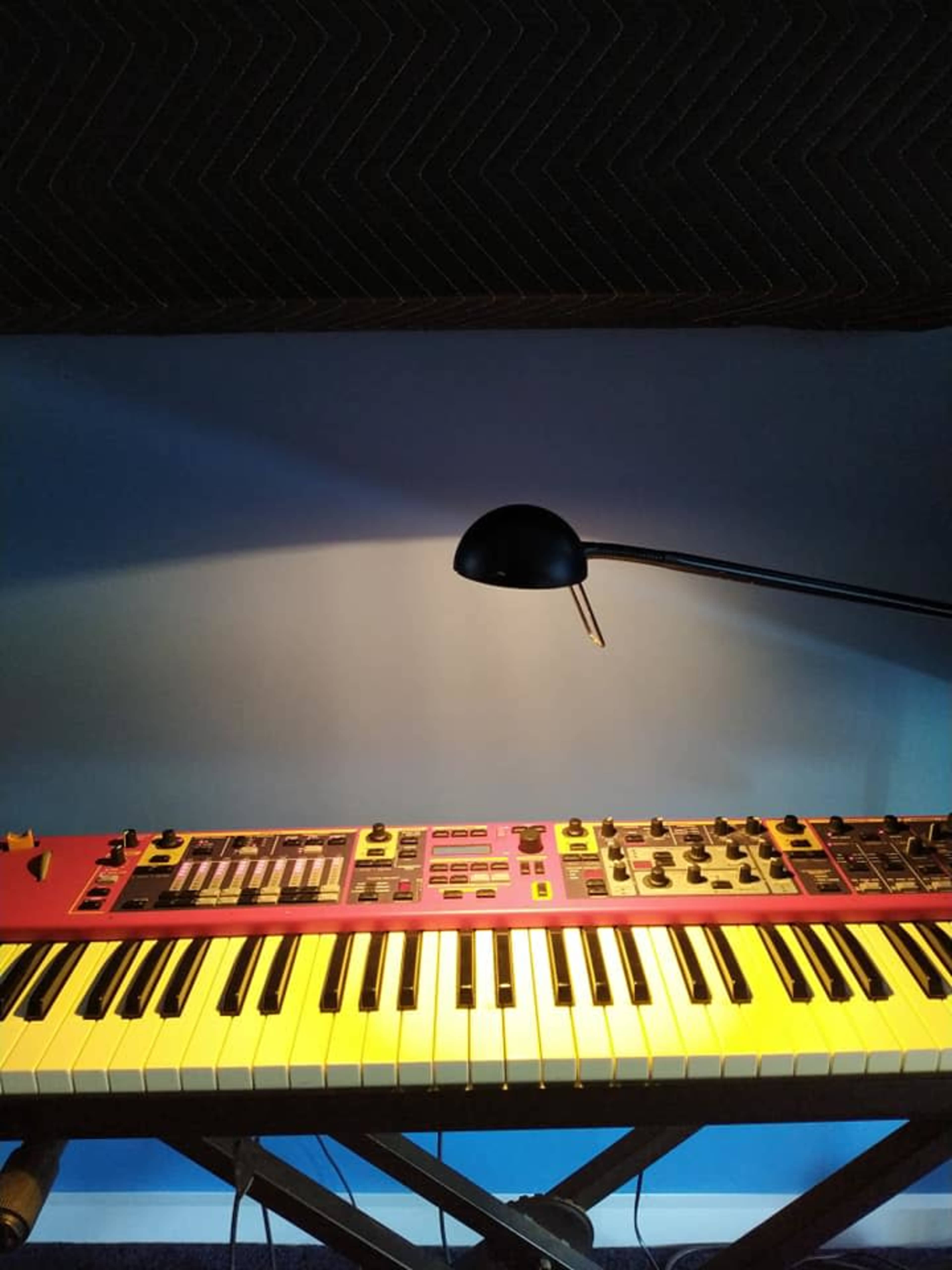 A synthesizer with a red panel and black keys sits on a wooden stand beneath a lamp, against a blue wall.