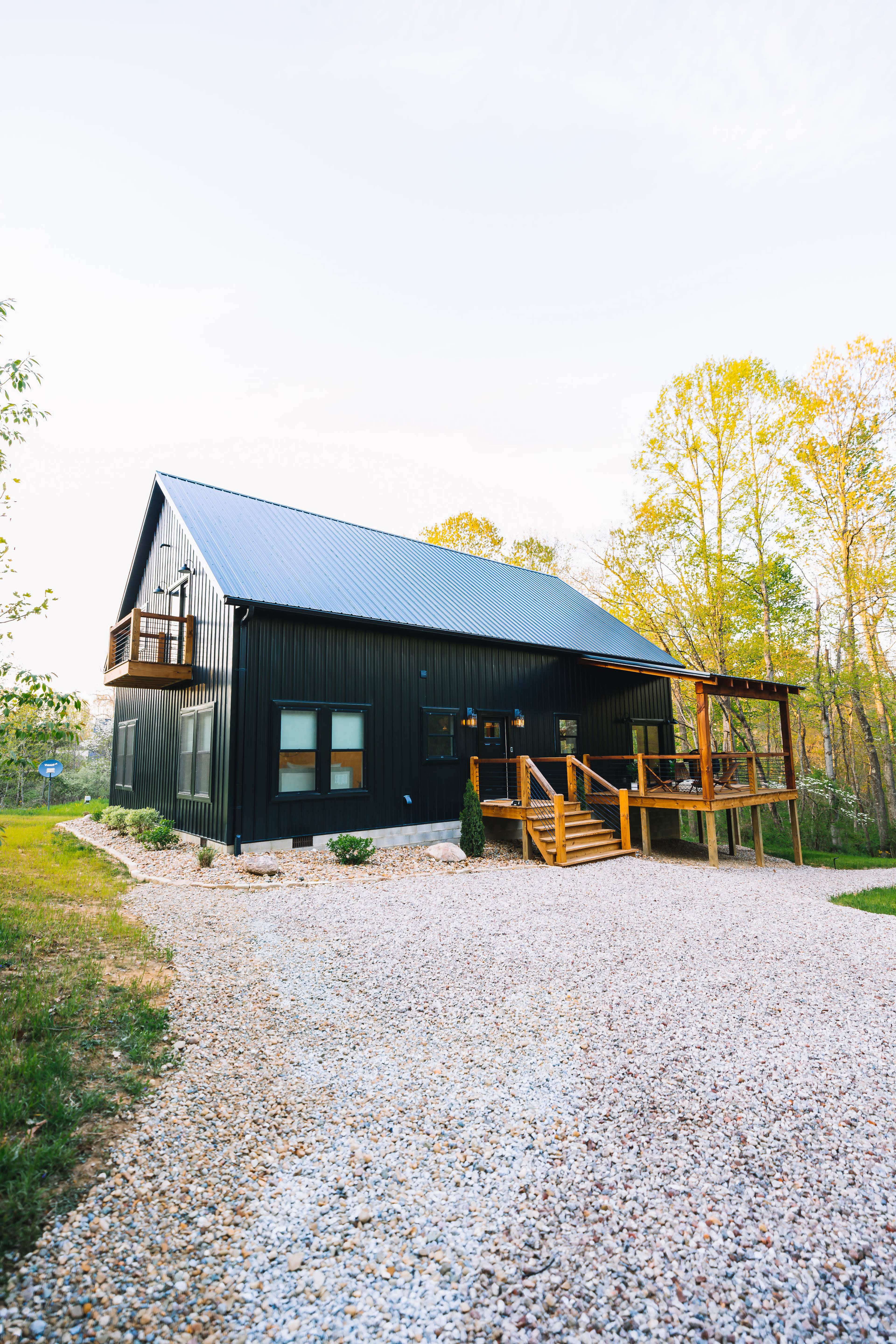A modern black metal house with a sloped roof and a wooden deck sits beside a gravel driveway, surrounded by trees.