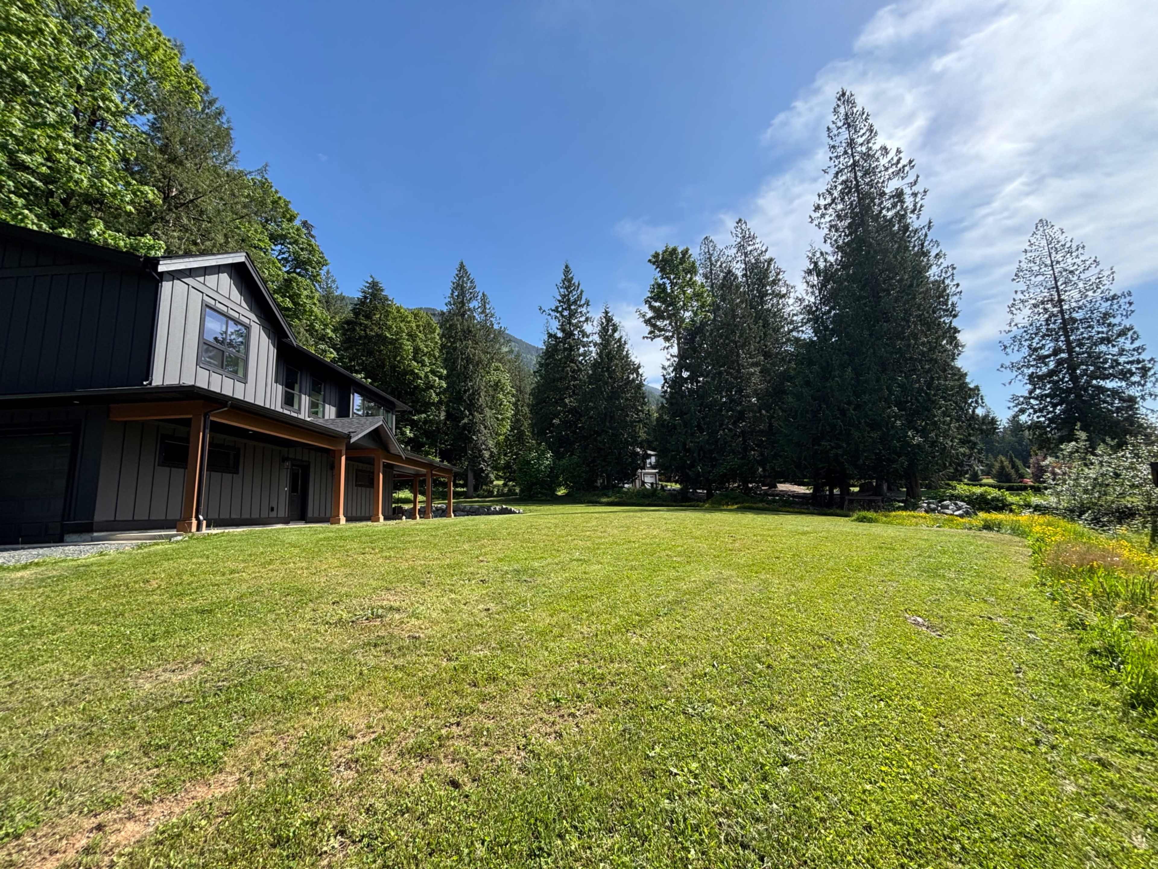 A modern house with large windows is situated on a spacious lawn surrounded by tall trees under a clear blue sky.