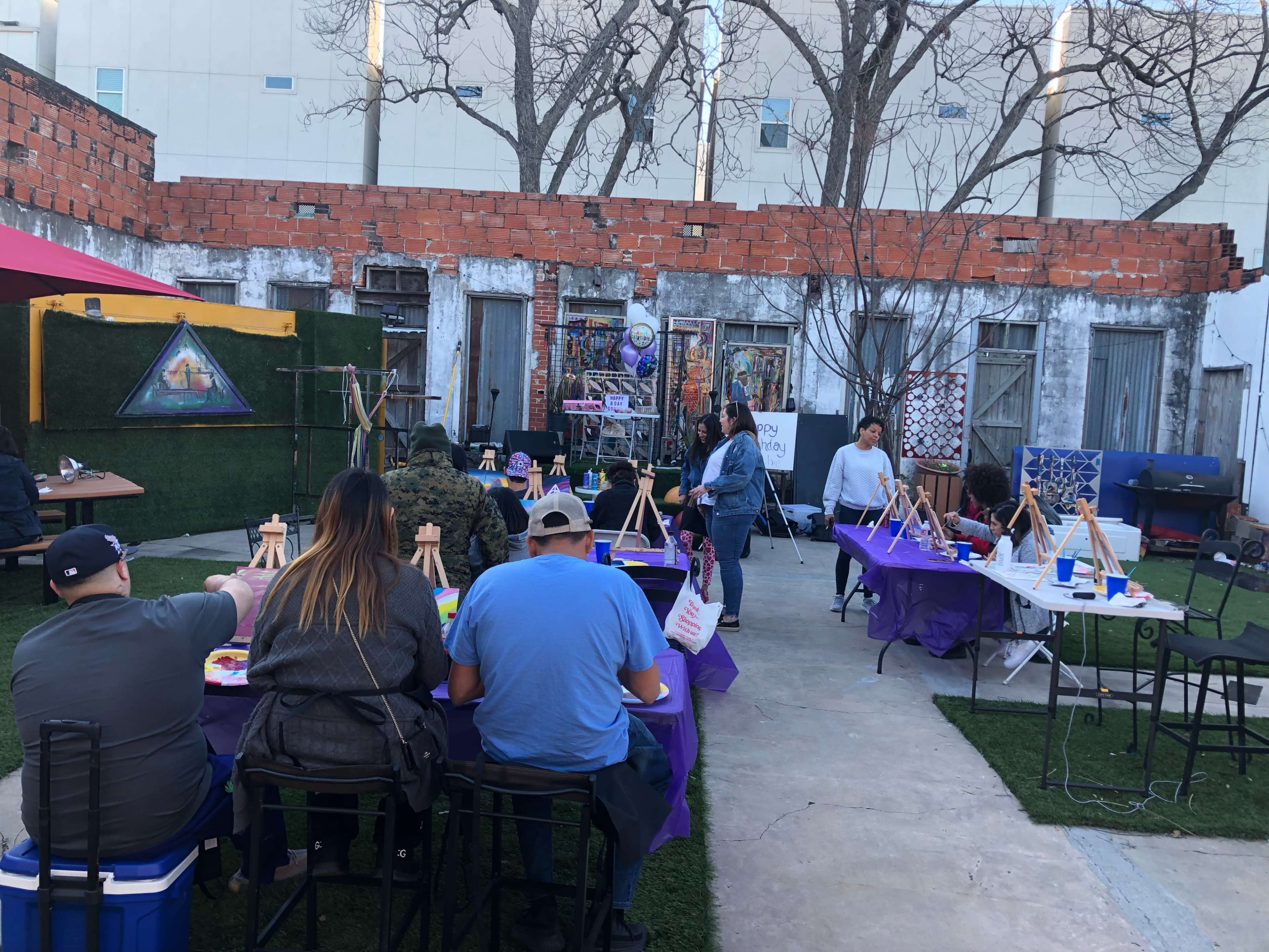 A group of people is seated at tables in an outdoor space, preparing to paint on easels.