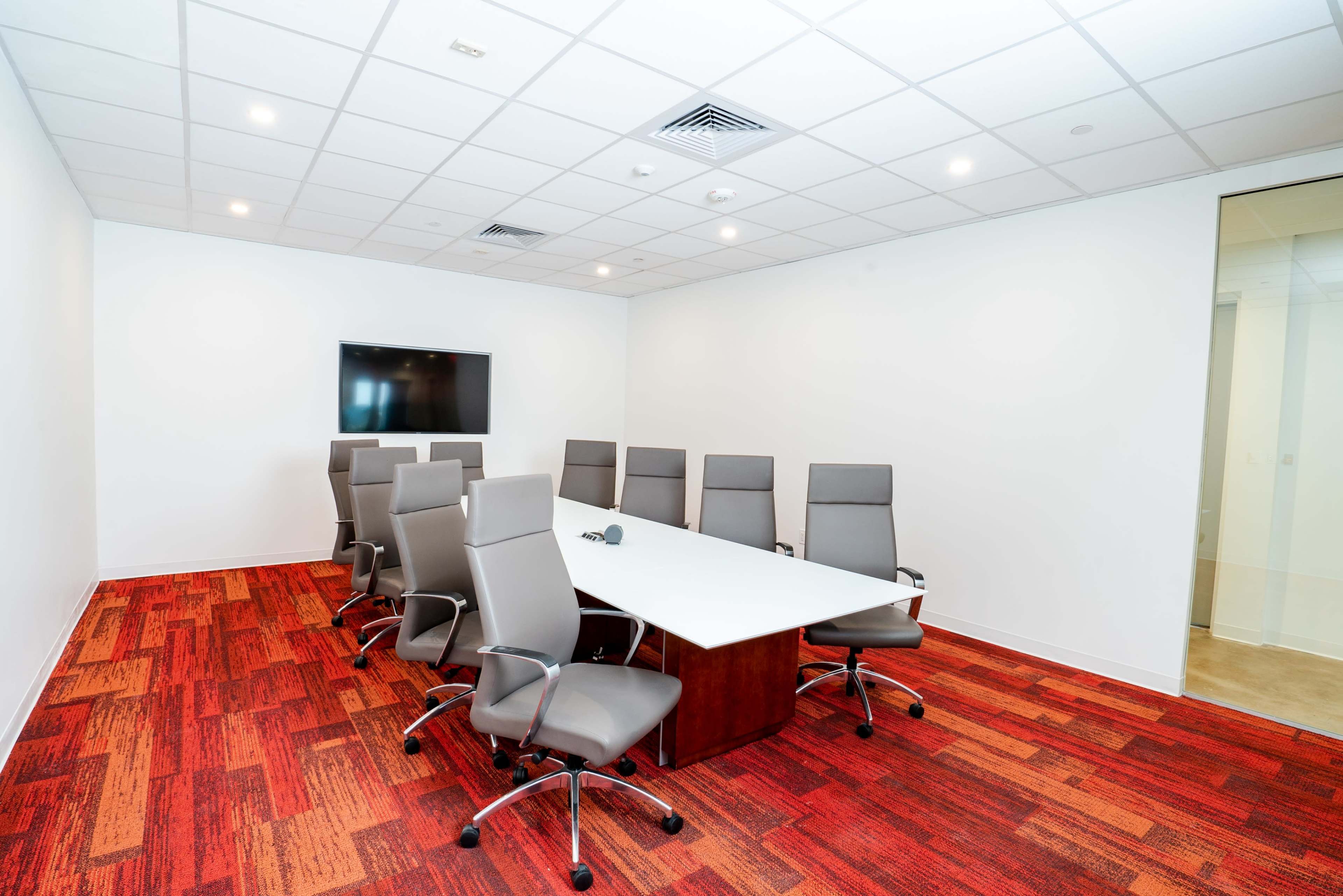 A modern conference room with a rectangular table surrounded by eight ergonomic chairs and a wall-mounted screen.