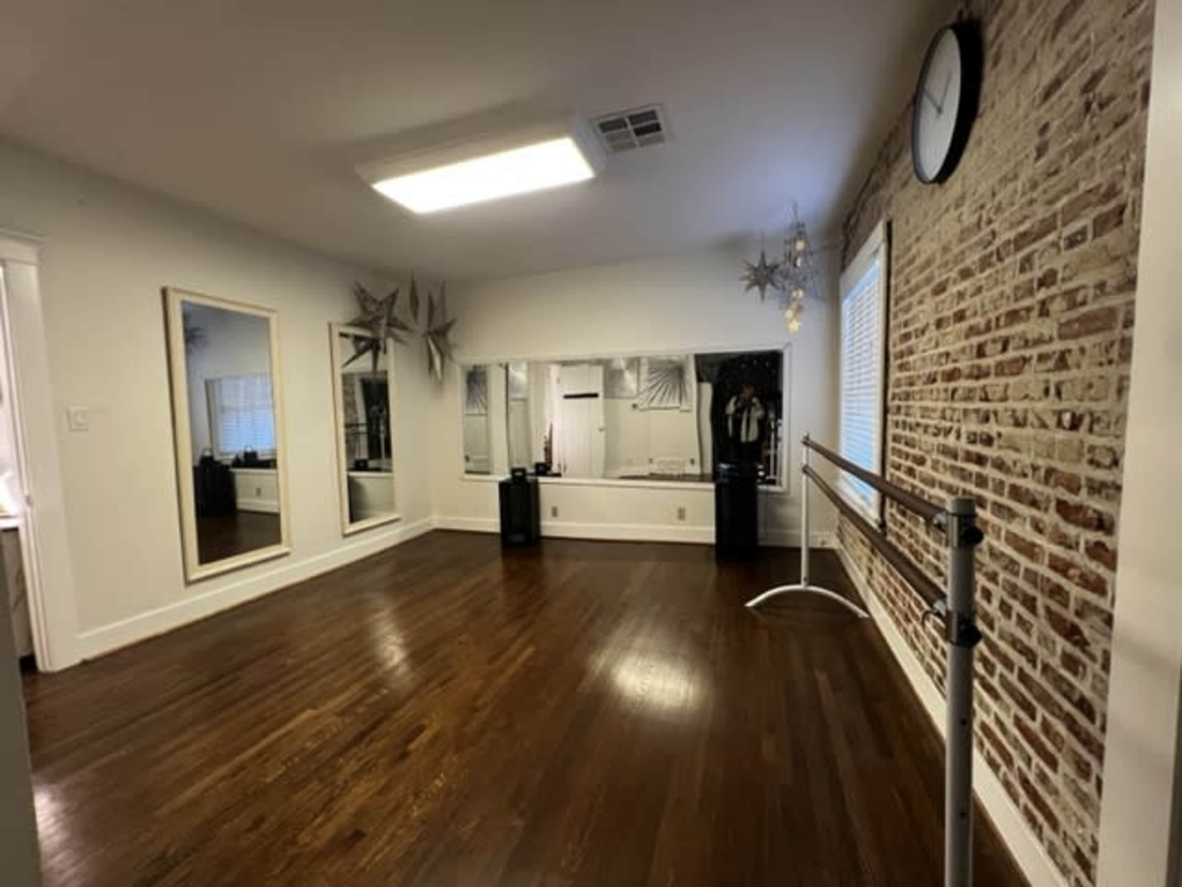 The room features hardwood floors, a brick wall, a large mirror, a ballet bar, and a clock on the wall, all illuminated by overhead lighting.