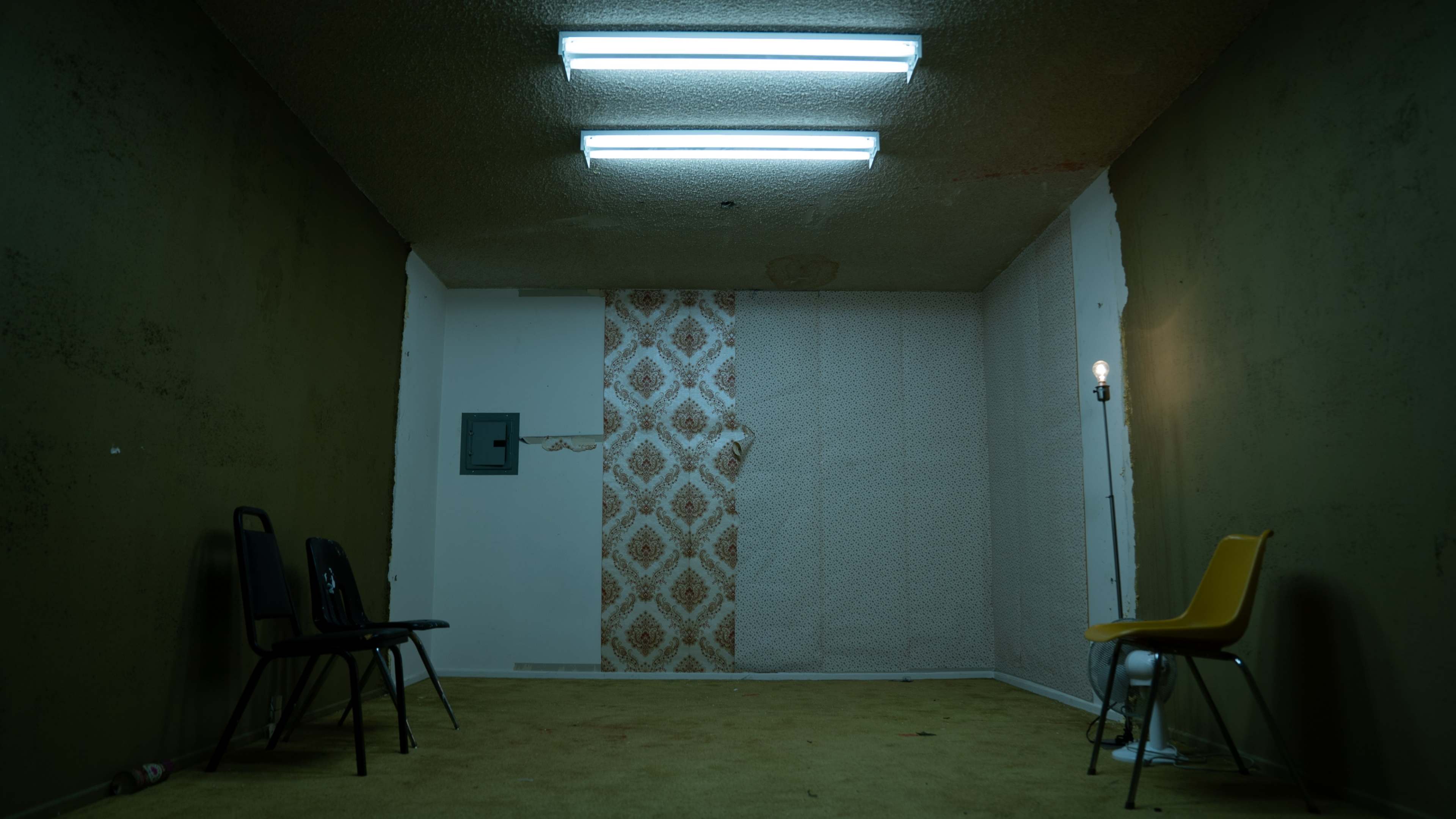 The image shows a sparse, empty room with two black chairs and one yellow chair, illuminated by fluorescent lights and featuring bare walls and carpet.