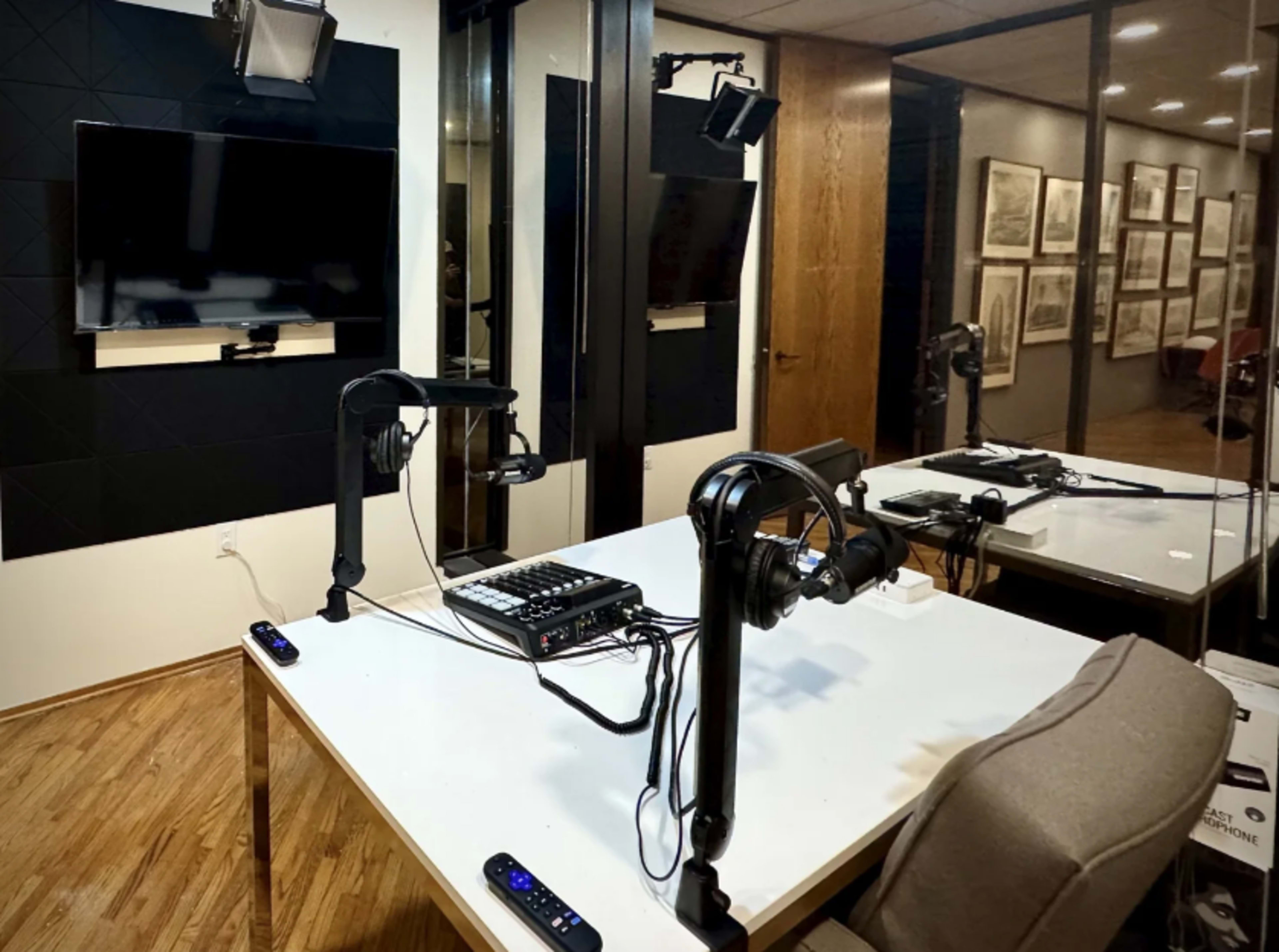The image shows a recording studio with a table equipped with microphones and audio equipment, situated in a glass-enclosed room.