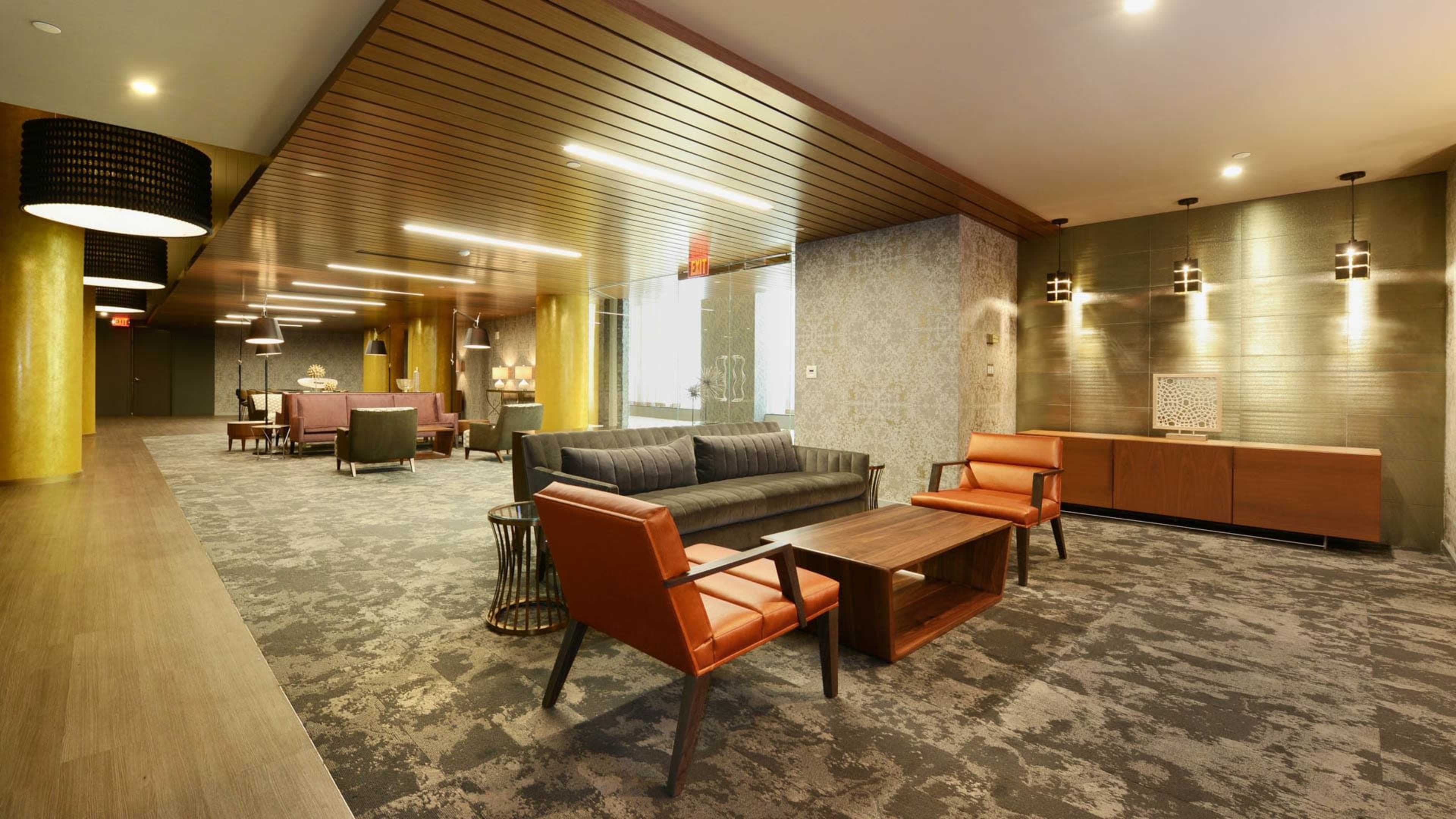A modern lounge area with a gray sofa, orange chairs, a wooden coffee table, and contemporary lighting fixtures along the walls.