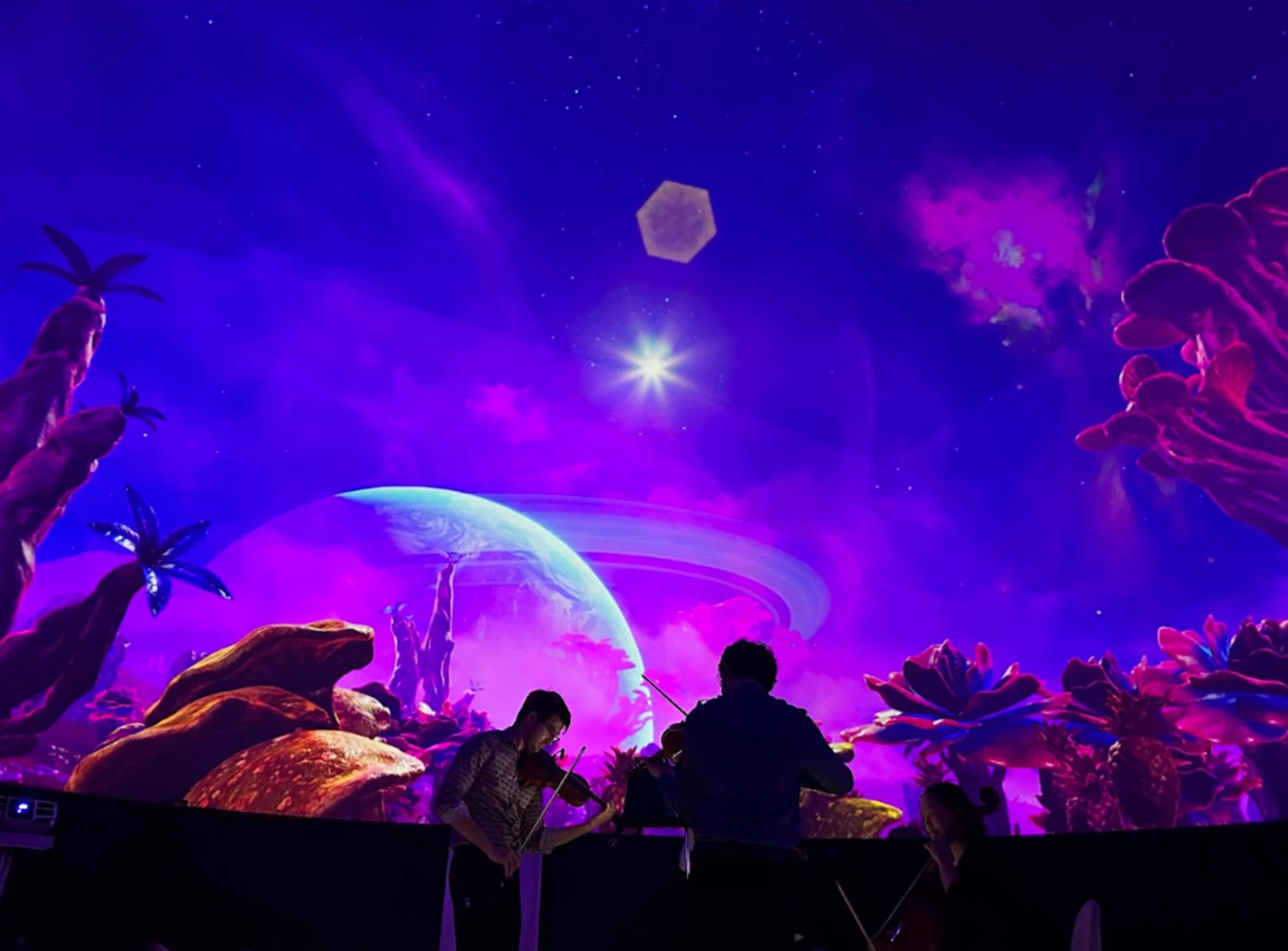 A group of musicians plays string instruments under a vividly colorful projection of an alien landscape with planets and stars.