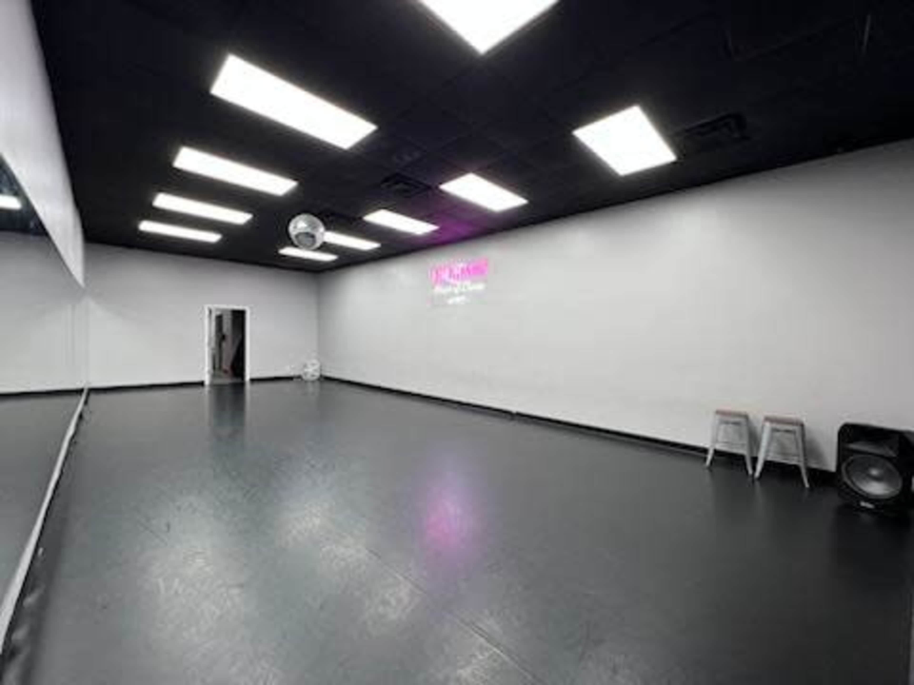 The image shows a spacious dance studio with black flooring, white walls, and bright overhead lights.