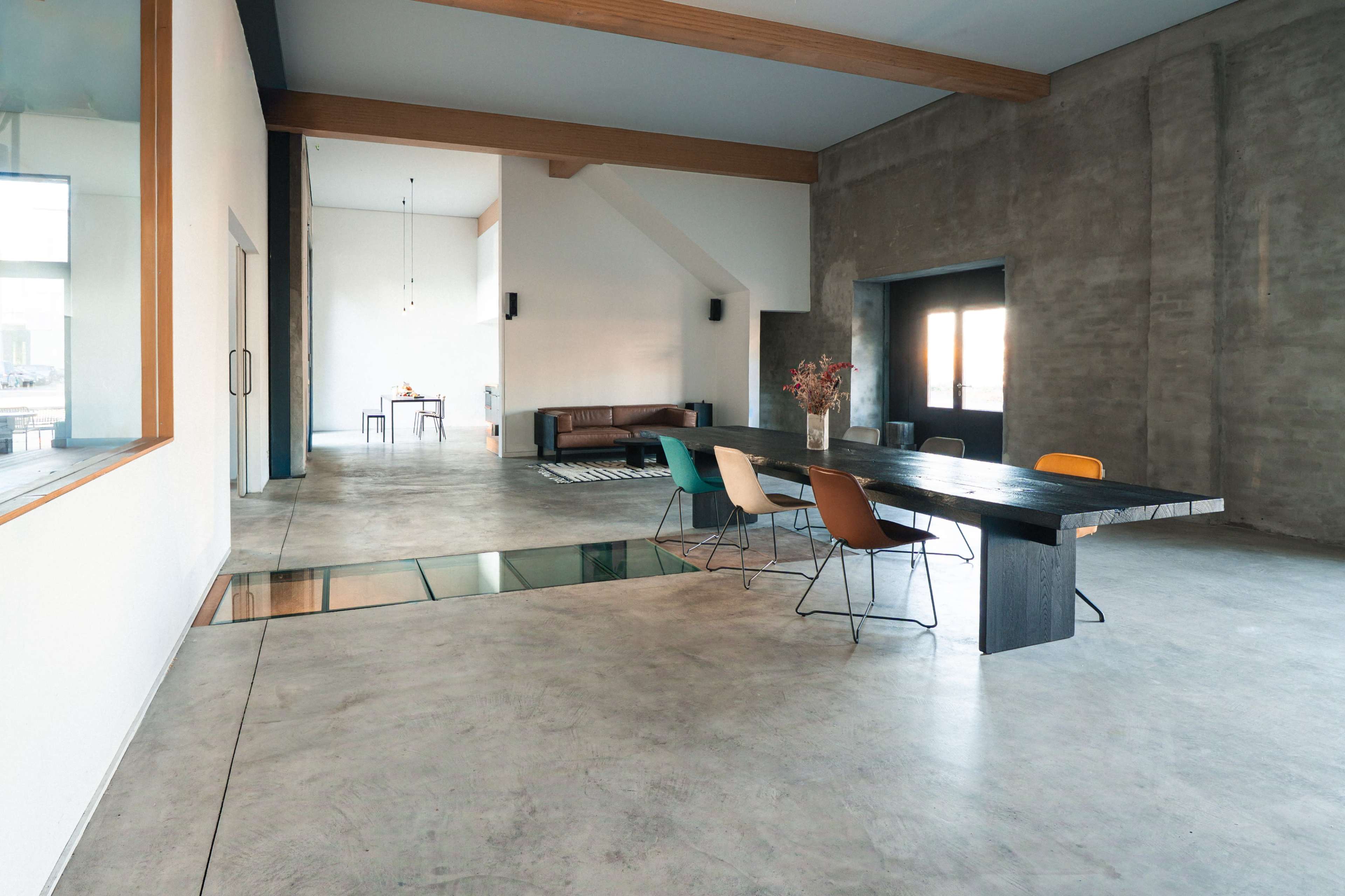 The interior features an open space with a concrete floor, wooden beams, a large dining table with colorful chairs, and a minimalist design.