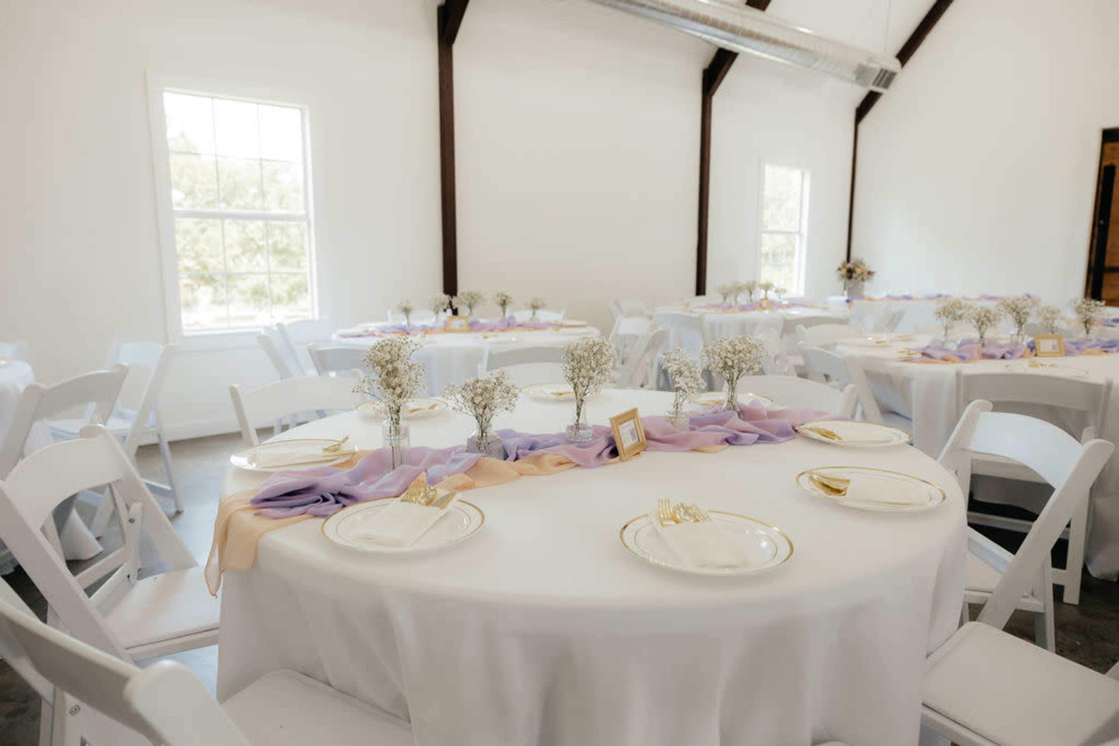 A reception hall with round tables covered in white tablecloths and adorned with purple and cream fabric runners, featuring small flower arrangements in glass vases.