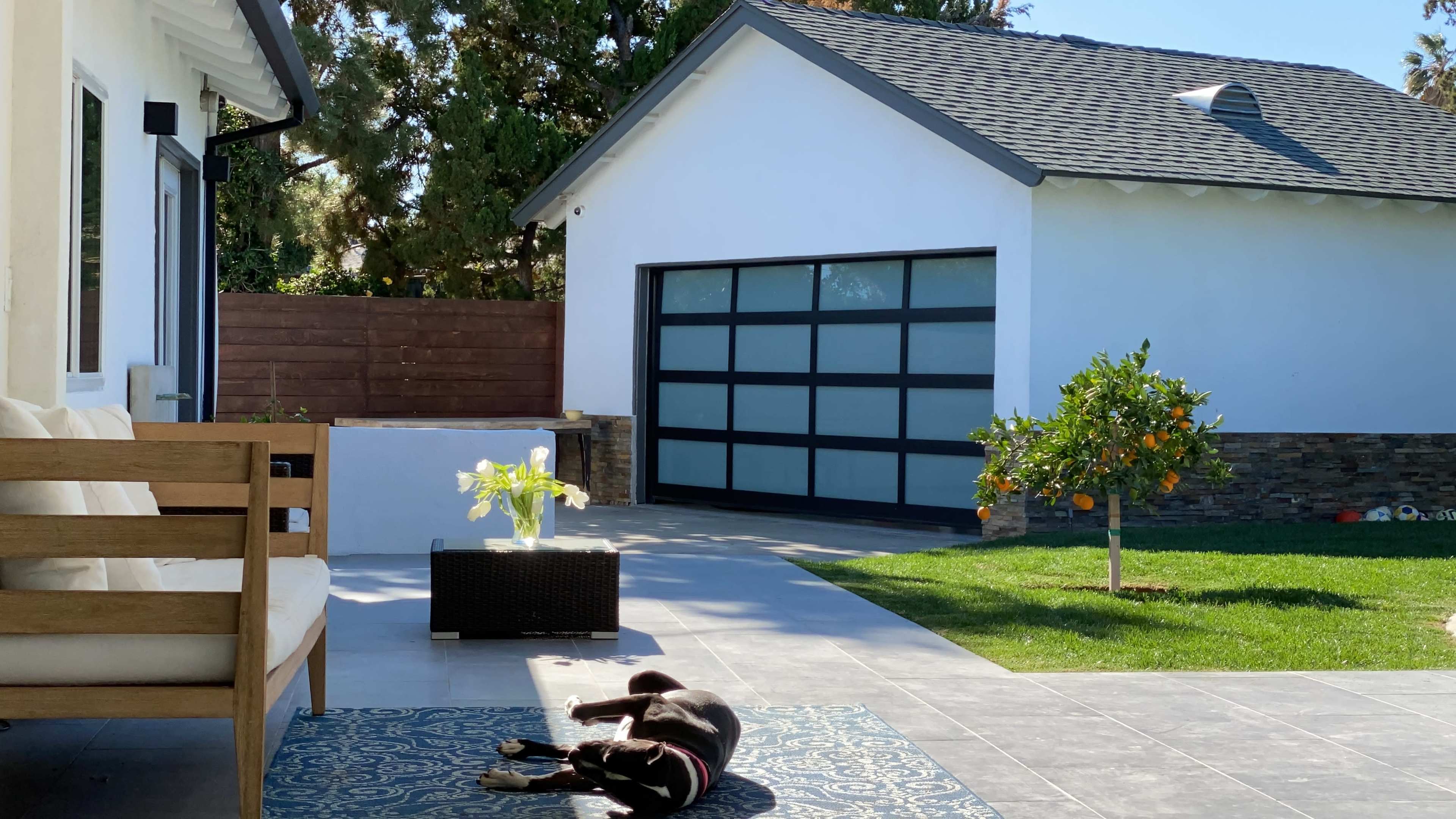 A black dog lies on a patterned rug in a sunny outdoor space in front of a modern home with a garage.