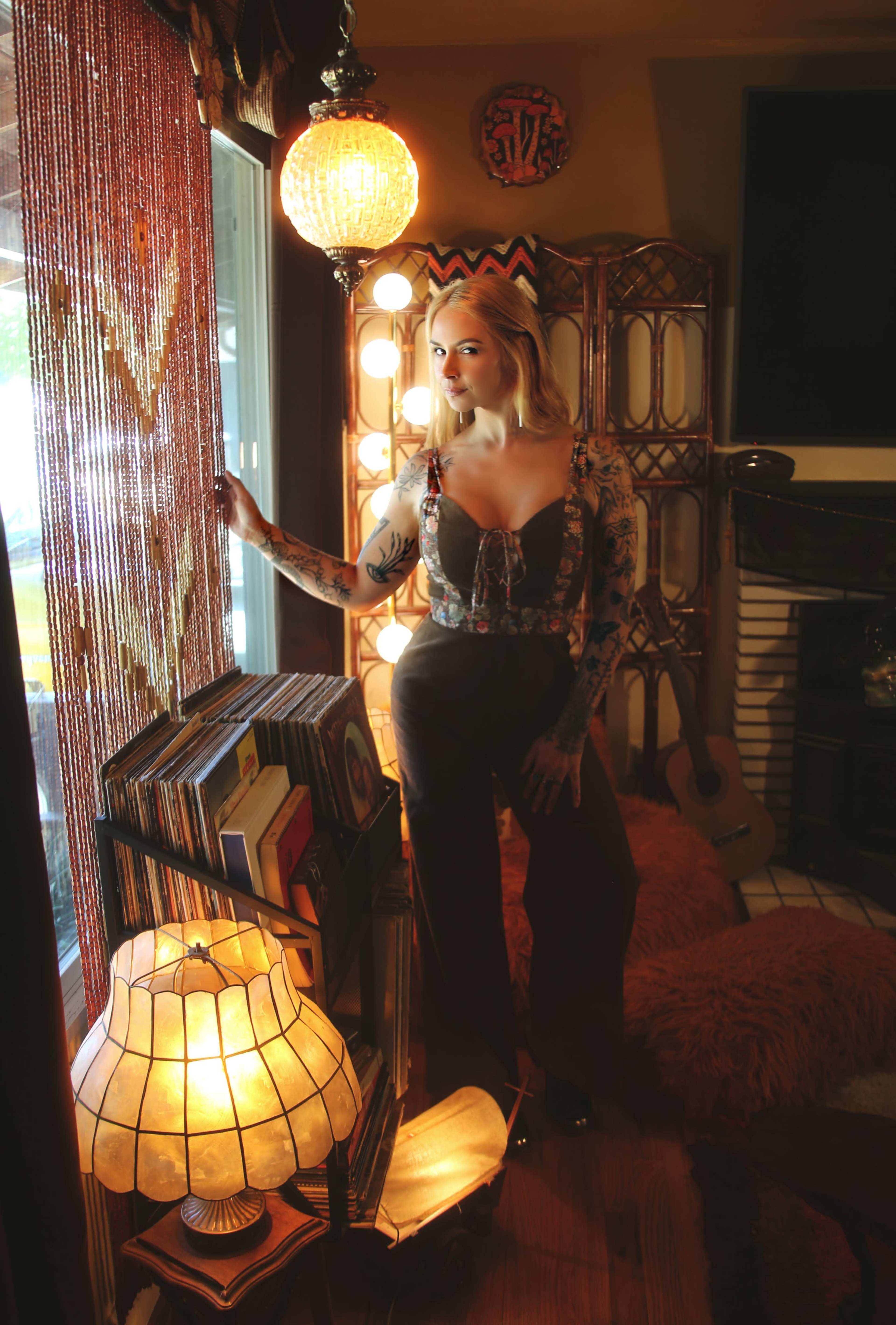 A woman with tattoos stands by a window in a warmly decorated room filled with books, a guitar, and artistic lighting.