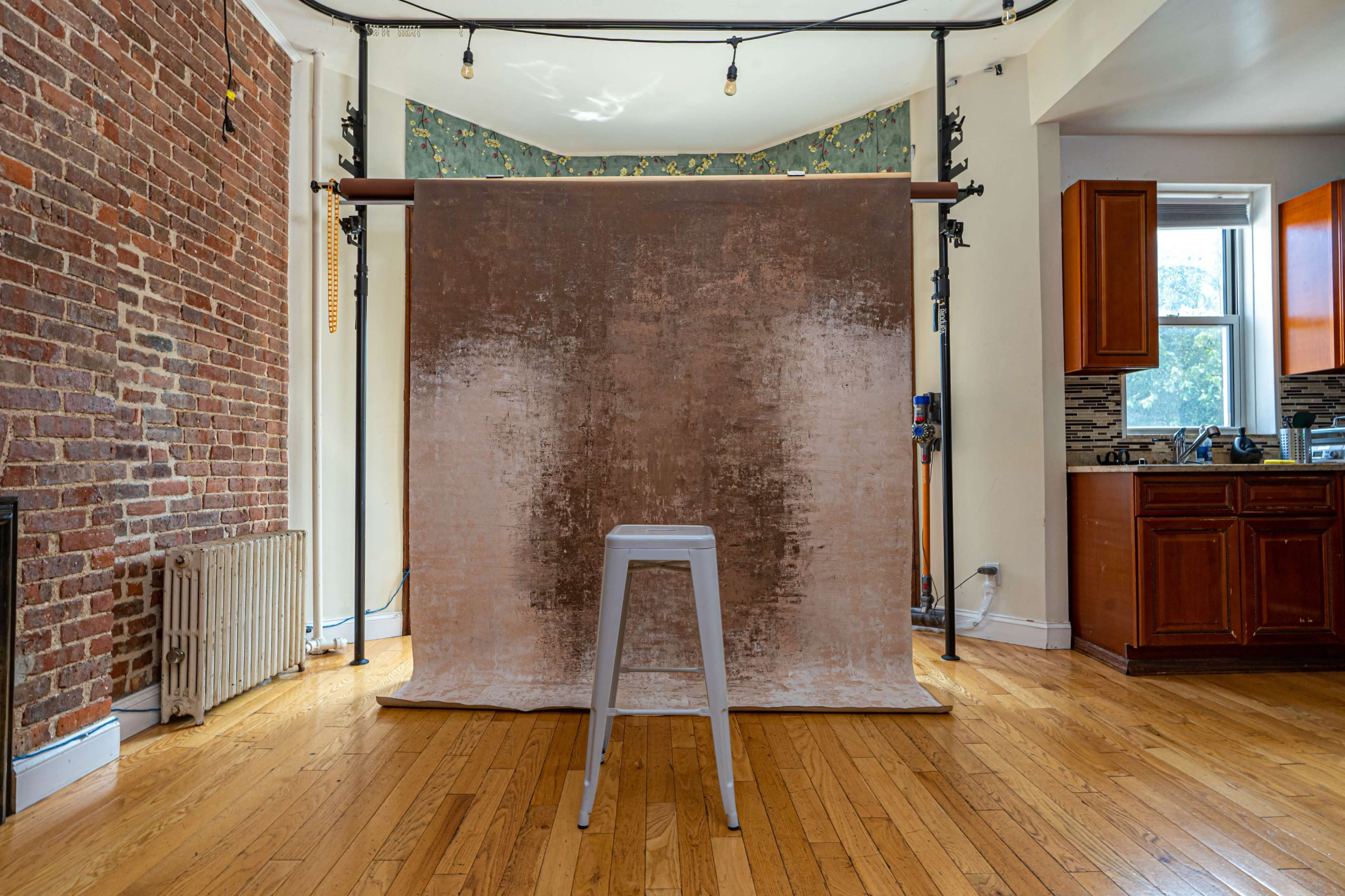 A photography studio setup with a brown backdrop, a metal stool in front, and a kitchen area visible in the background.