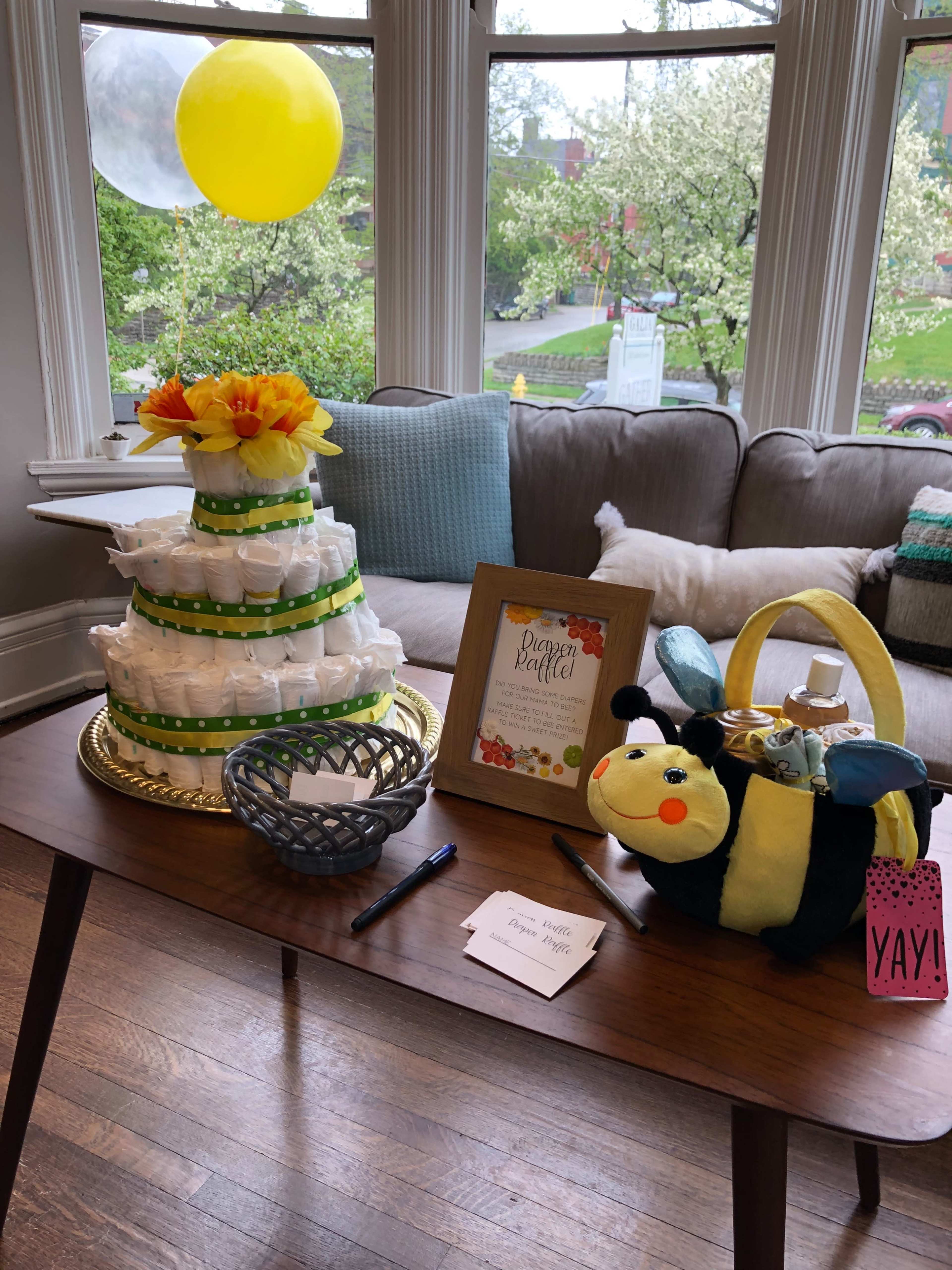 A multi-tiered cake decorated with yellow flowers is displayed on a table alongside a plush bee toy and a framed sign, in a well-lit room with large windows.