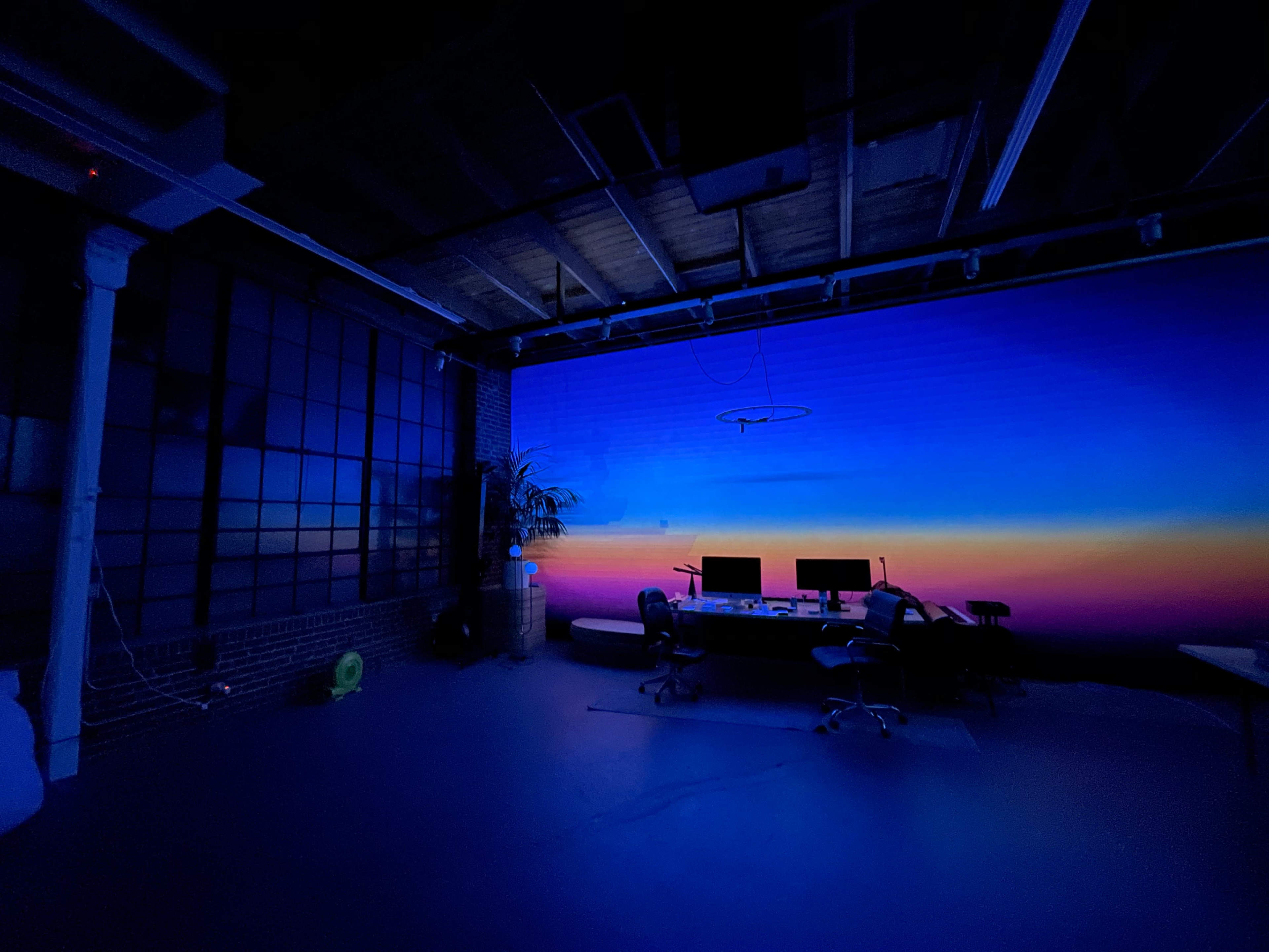 A dimly lit studio features a large wall with a gradient sunset backdrop and a conference table with several chairs arranged around it.