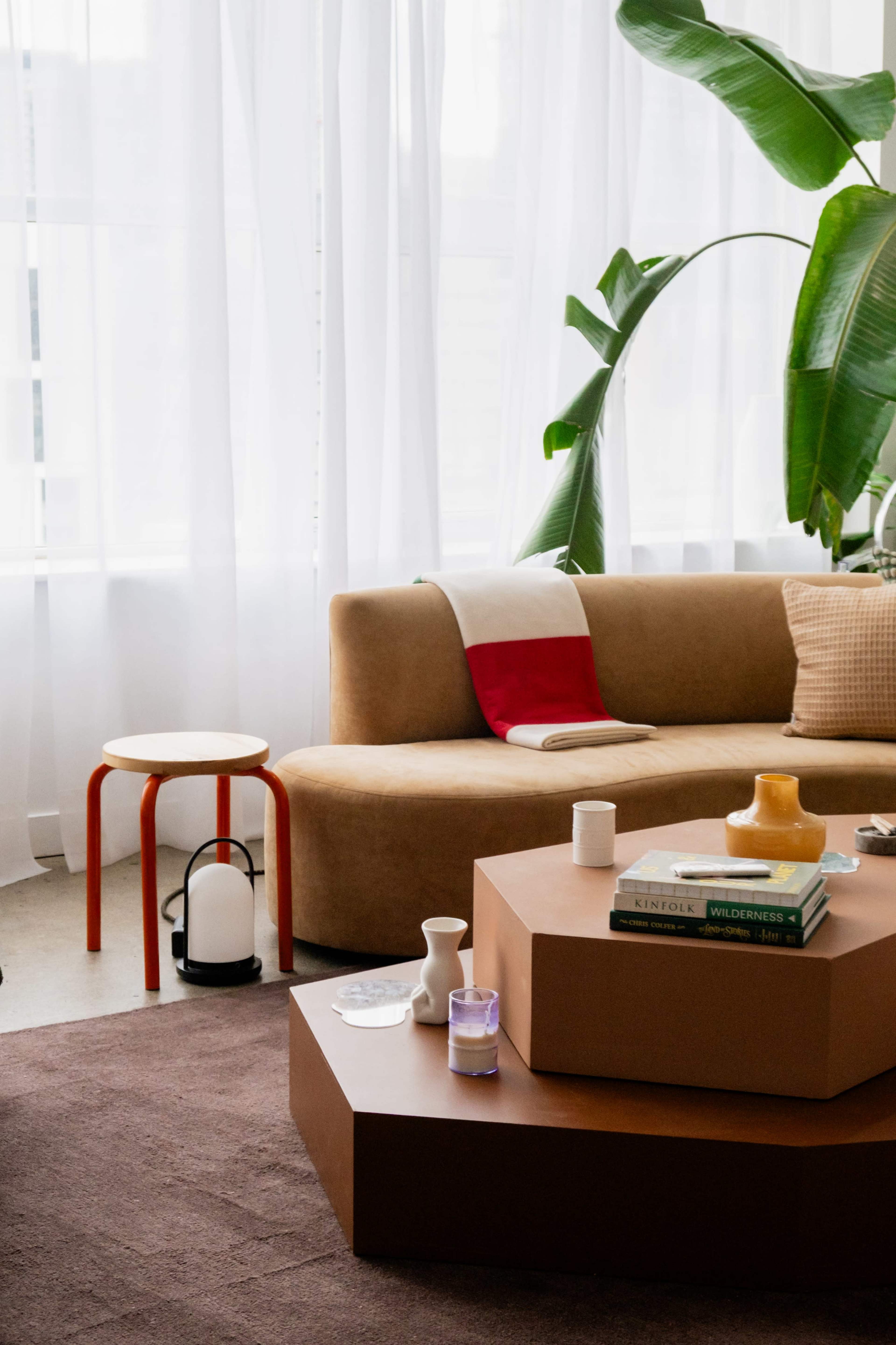 The image shows a cozy living room with a curved brown sofa, a small red side table, a stack of books on a geometric coffee table, and a large green plant near a window covered by sheer curtains.