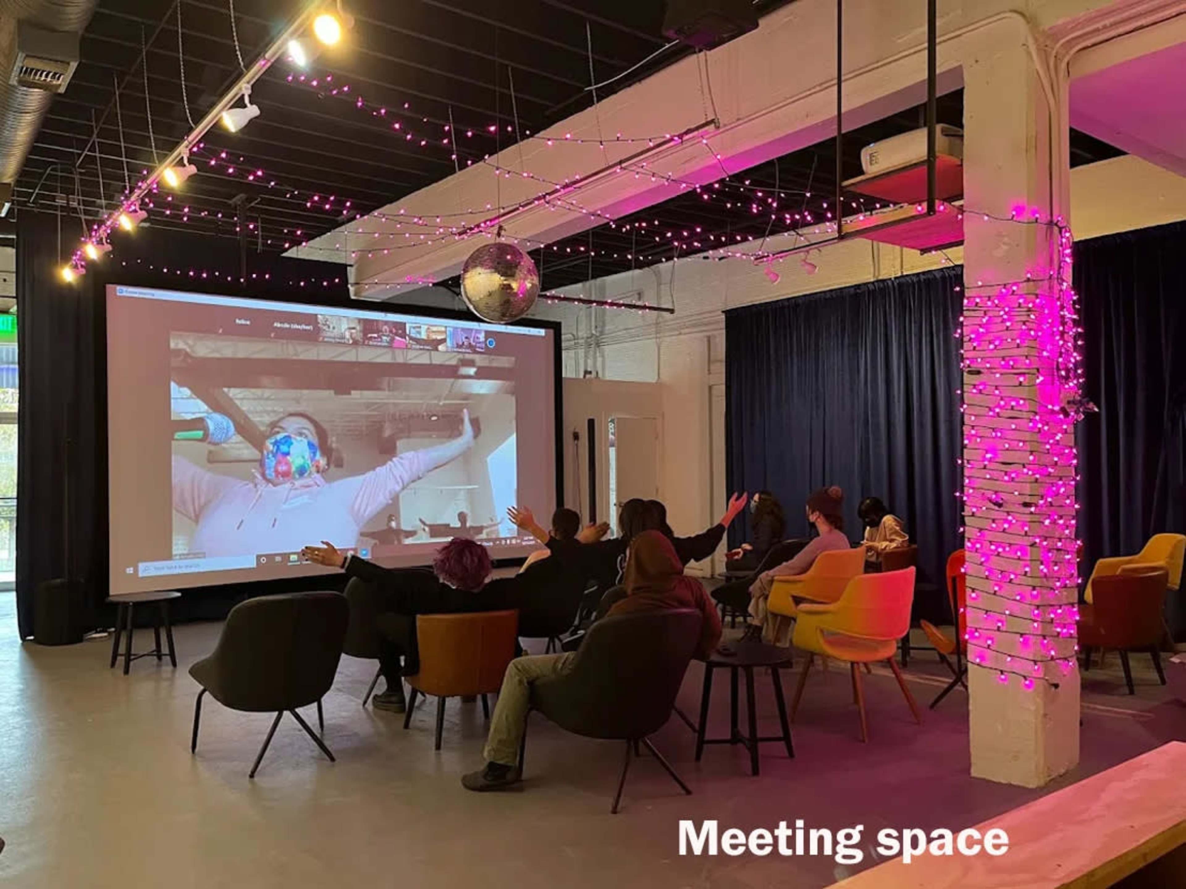 A group of people sits in a meeting space while participating in a video call displayed on a large screen.