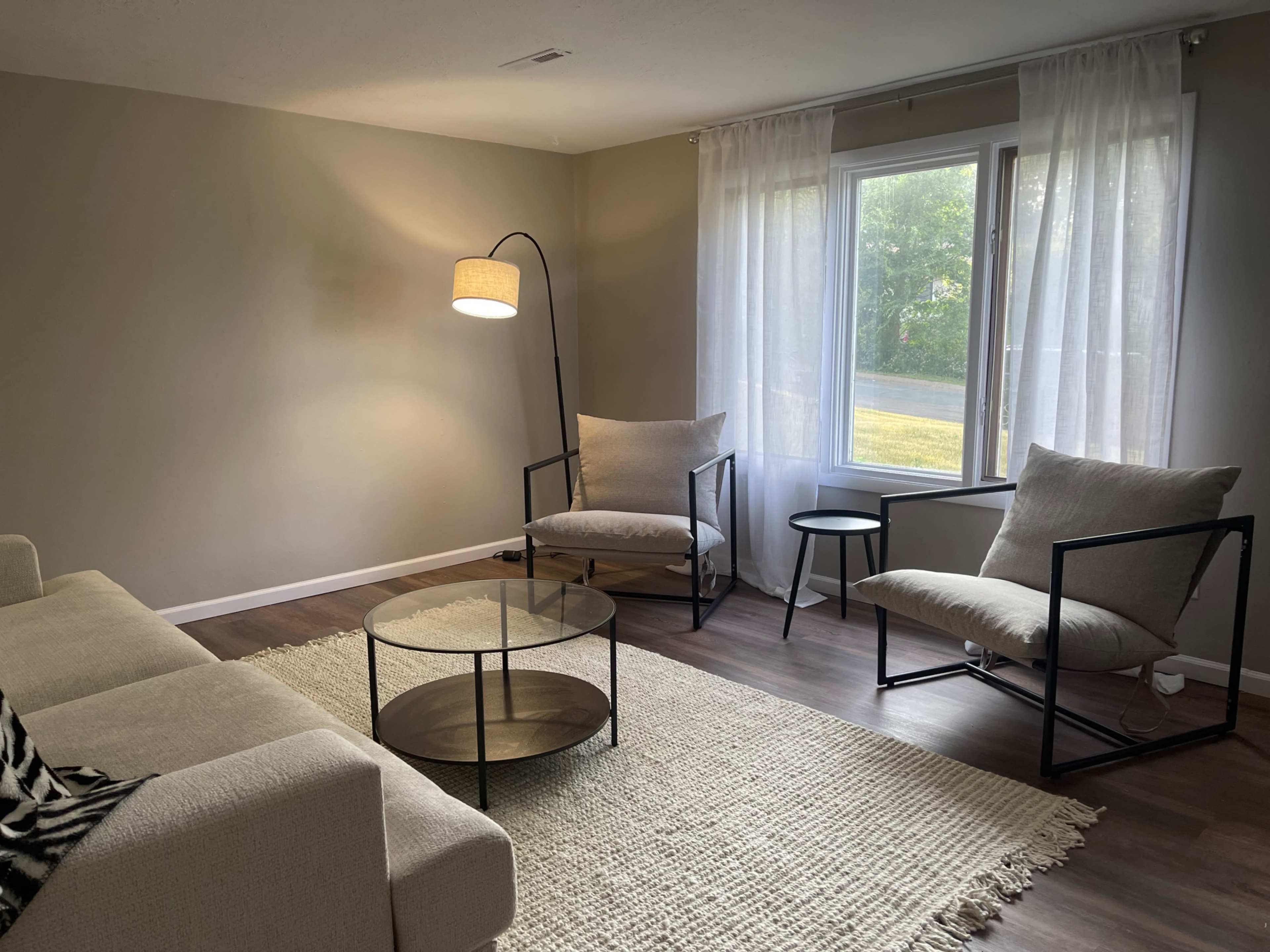 A cozy living room features two armchairs and a sofa arranged around a round coffee table, with a large window allowing natural light to enter.