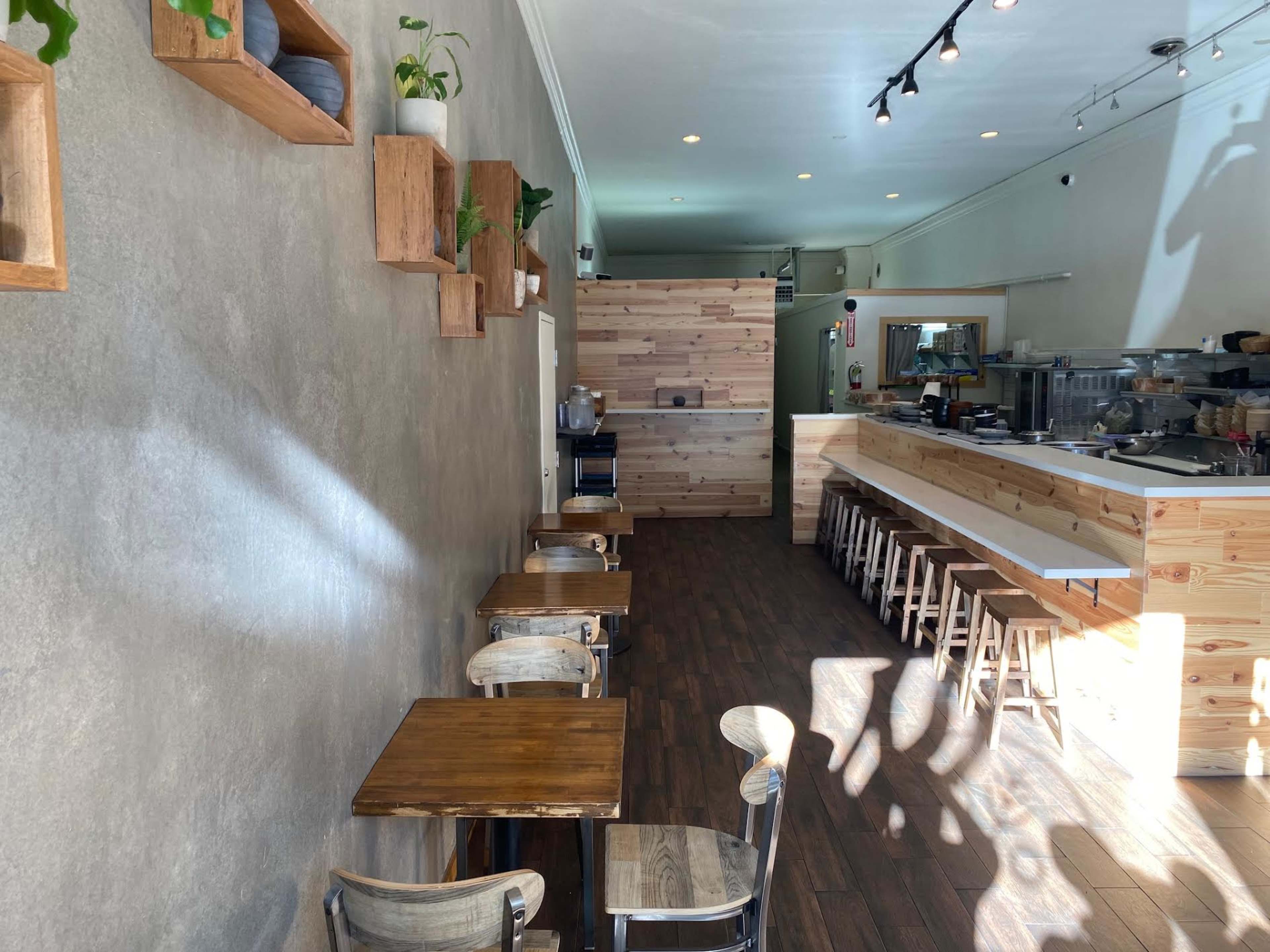 A minimalist café interior featuring wooden tables and chairs, a light-colored wall with plants on shelves, and a wooden counter in the background.