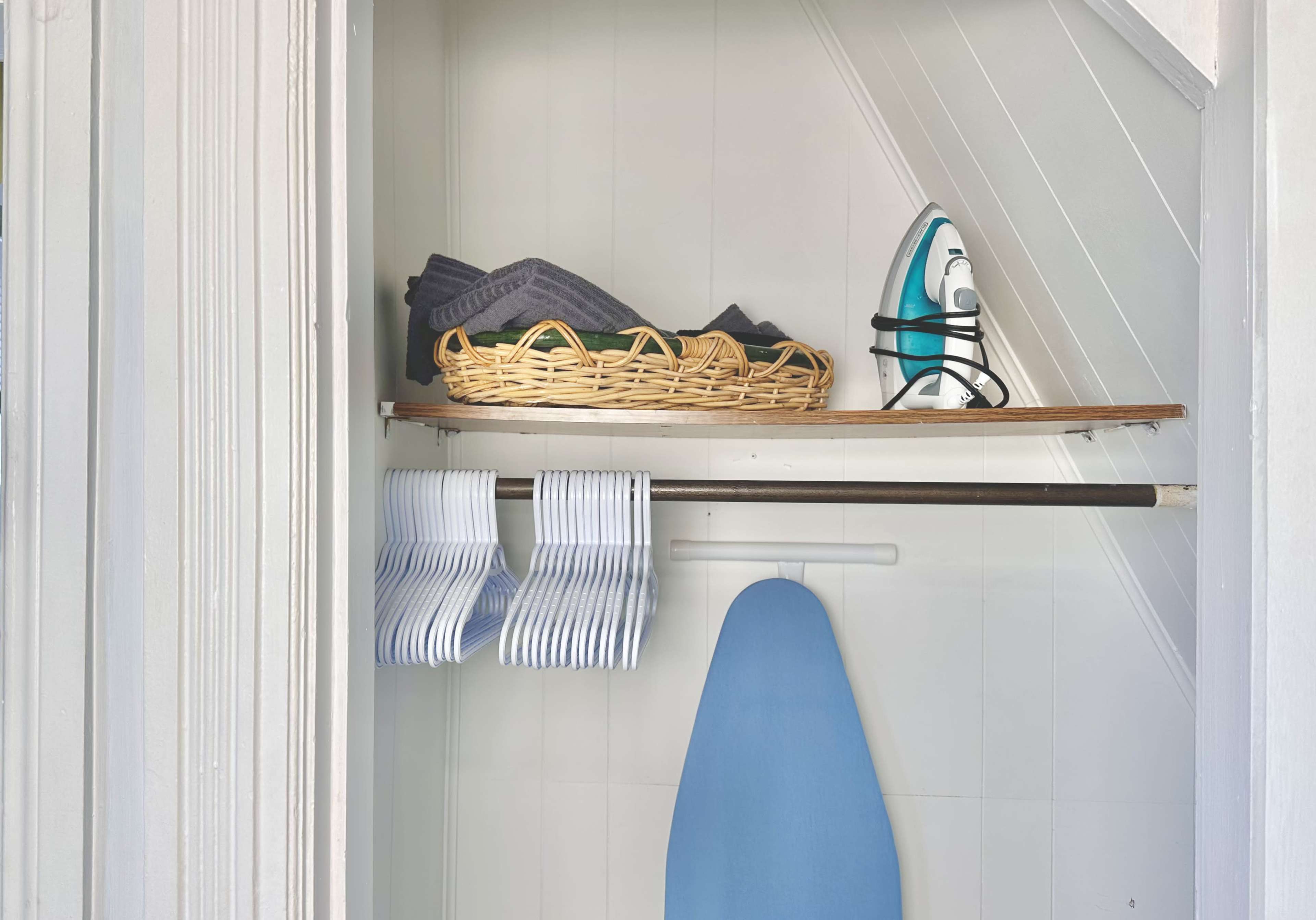 The image shows a small closet containing an ironing board, an iron, folded towels in a basket, and several neatly hung striped cloths on a rack.